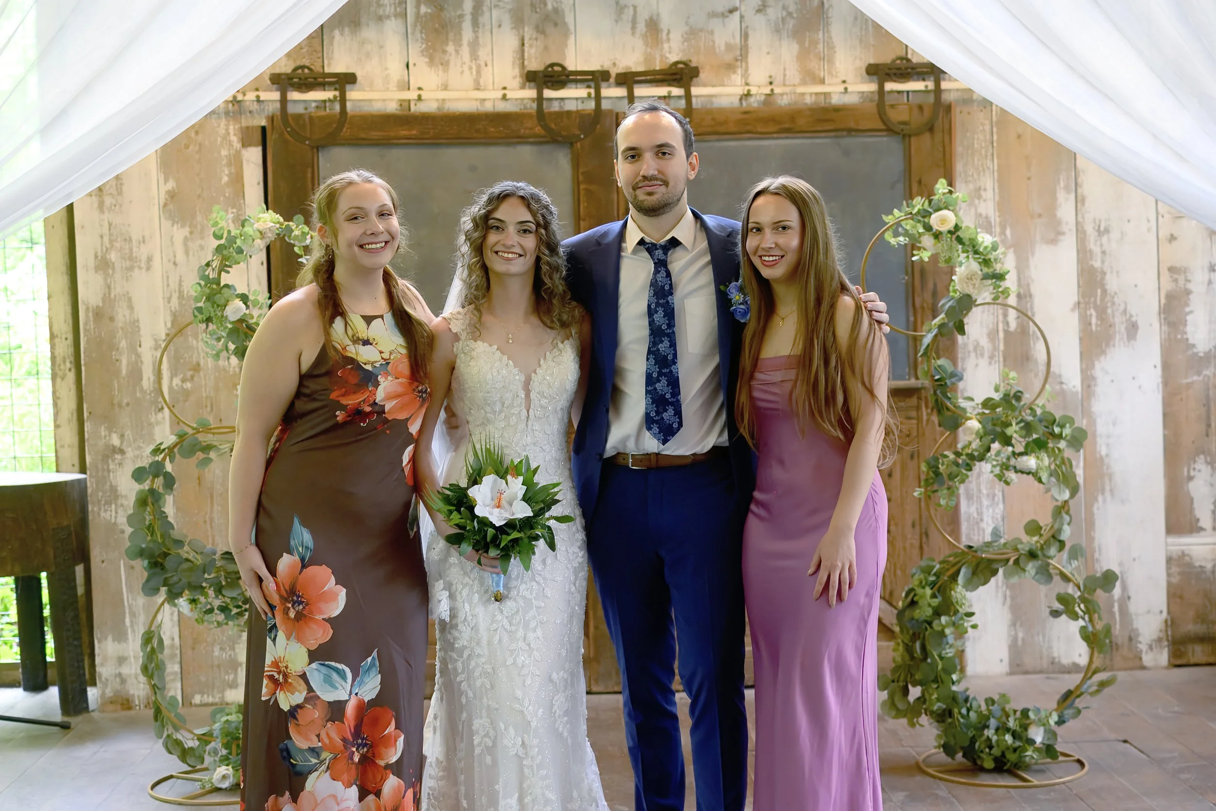 A wedding party of four people standing under a white draped canopy, with a wooden wall background and greenery decor, including a bride in a white lace dress holding a bouquet, and three women in dresses beside her, with a man in a suit and tie.