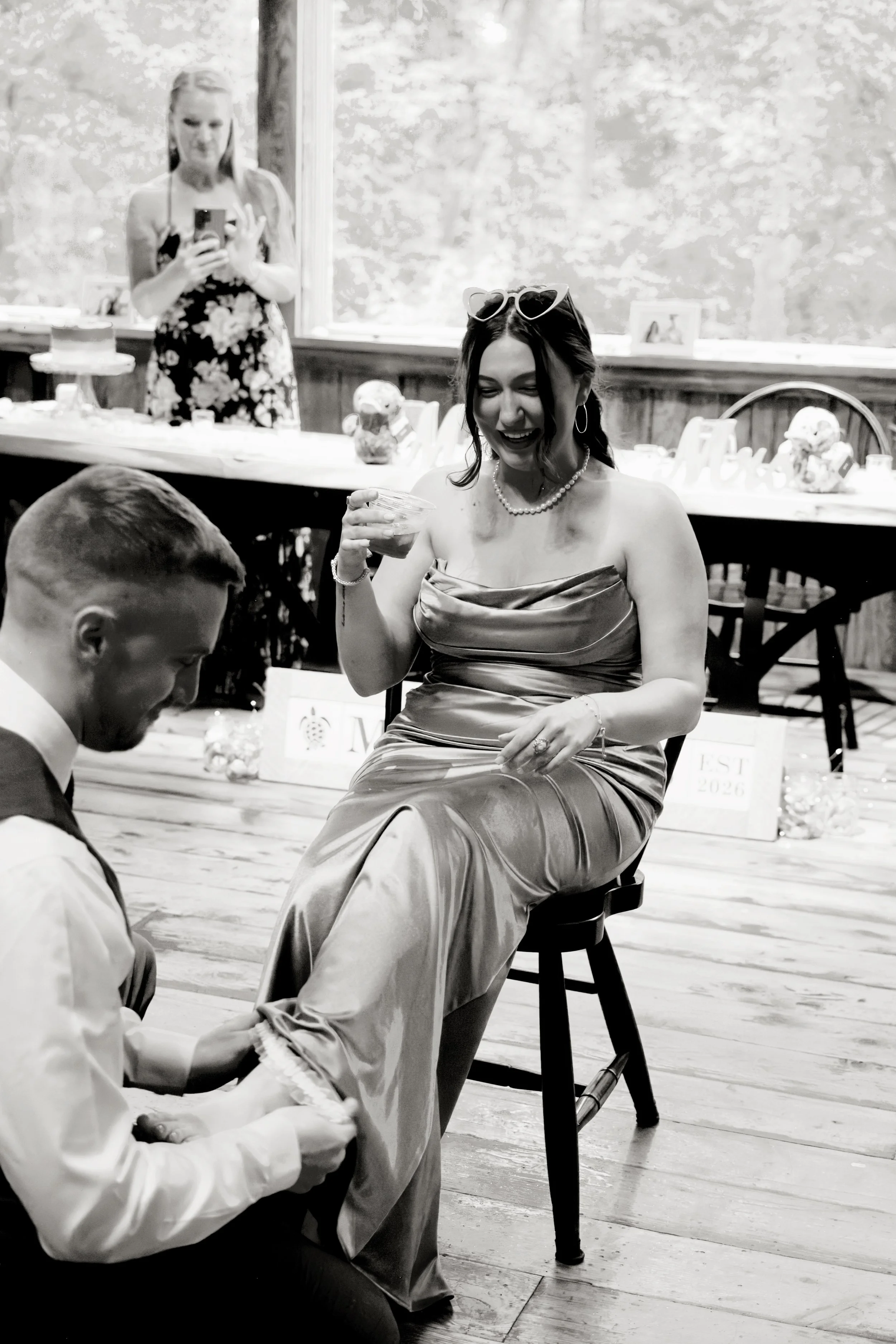 A woman in a strapless dress with sunglasses on her head, wearing pearls, is sitting on a chair laughing, holding a drink. There is a man in a white shirt sitting in front of her. In the background, a woman in a floral dress is taking a photo or vide