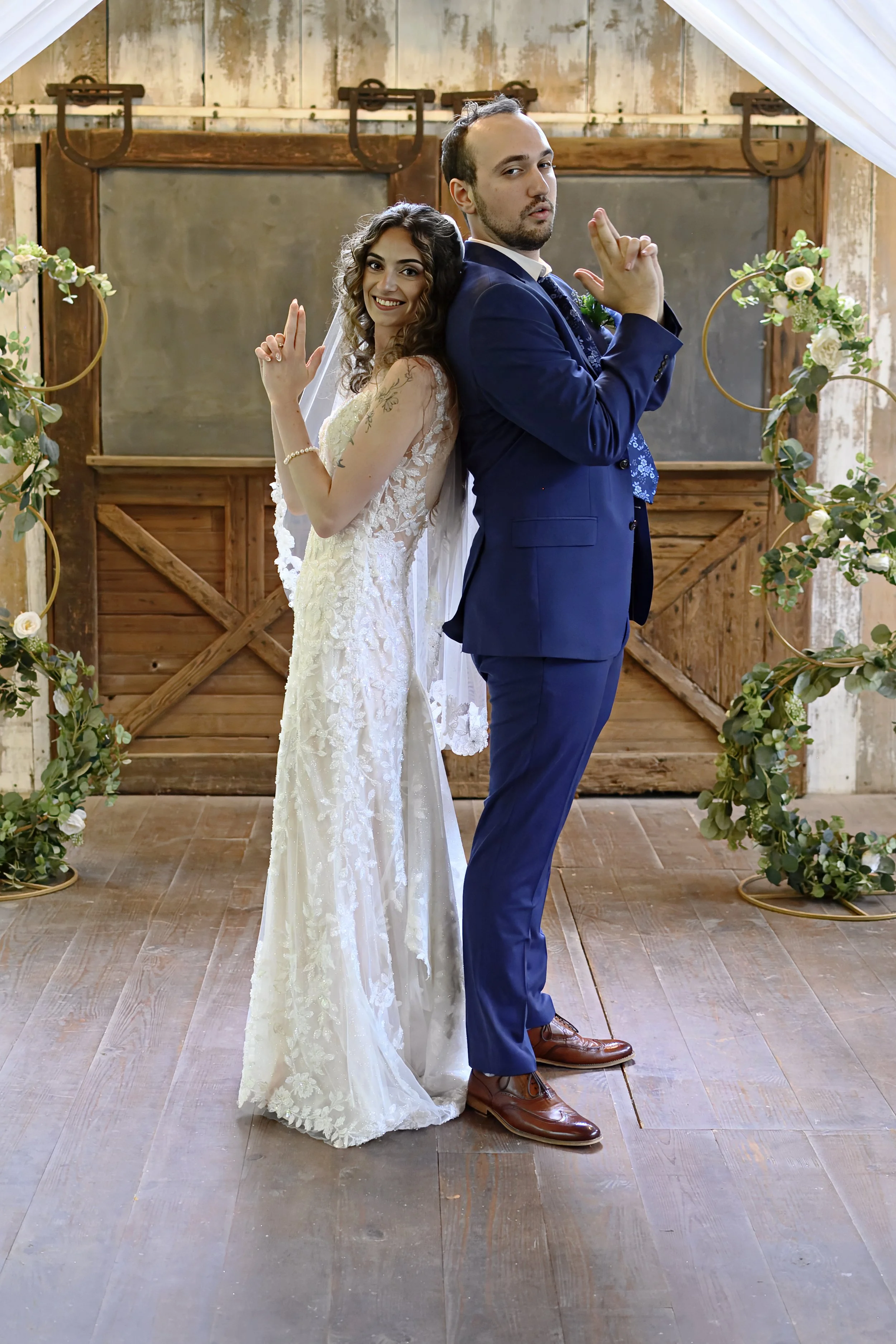 A bride and groom standing back-to-back in a rustic wedding setting, with the bride smiling at the camera and the groom with a serious expression. They are membuat hand gestures that resemble guns pointed outward, and are surrounded by floral arches.