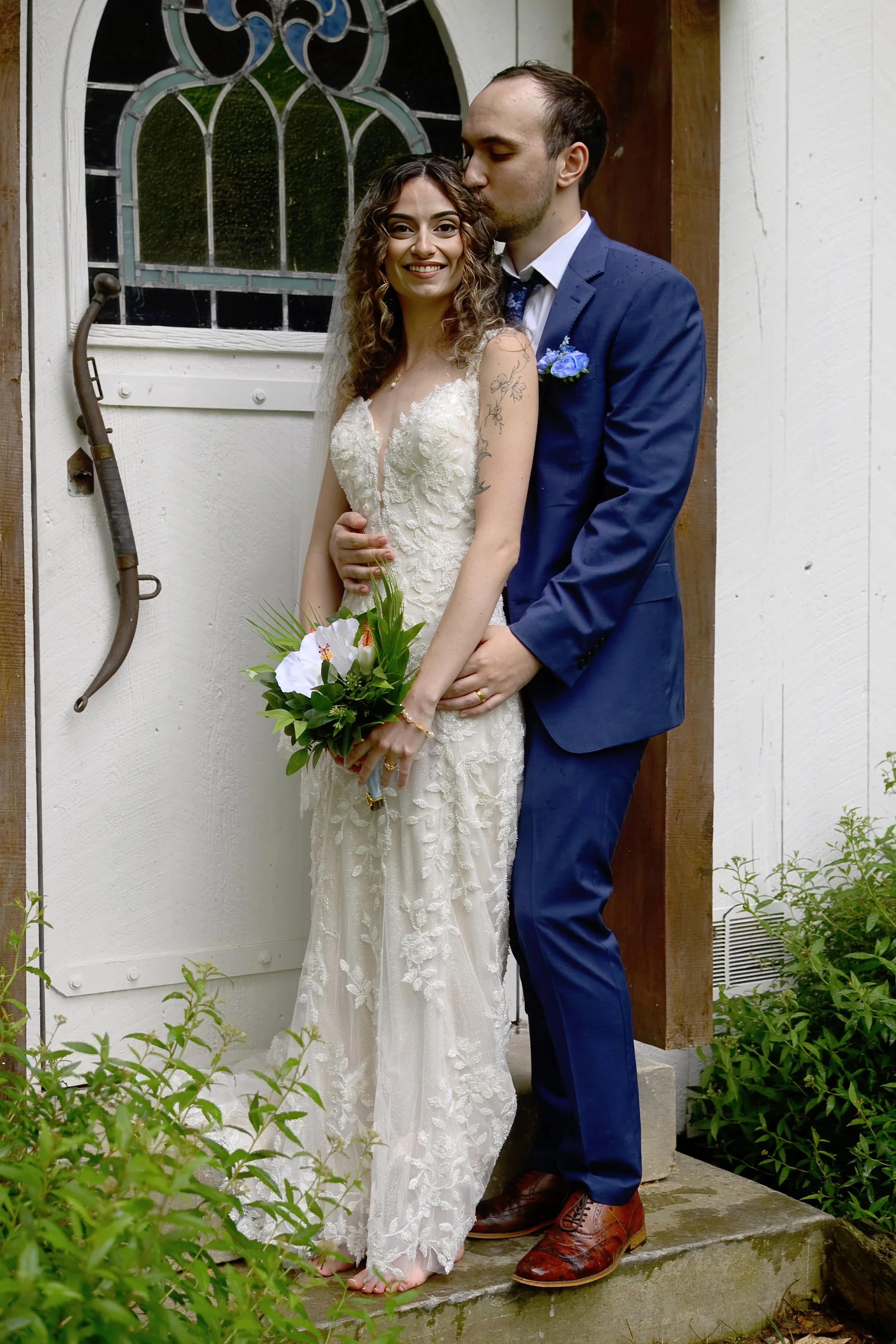 A bride in a lace wedding gown holding a bouquet of white lilies and greenery, standing next to a groom in a navy blue suit, outdoors near a white wall with a stained glass window and greenery.