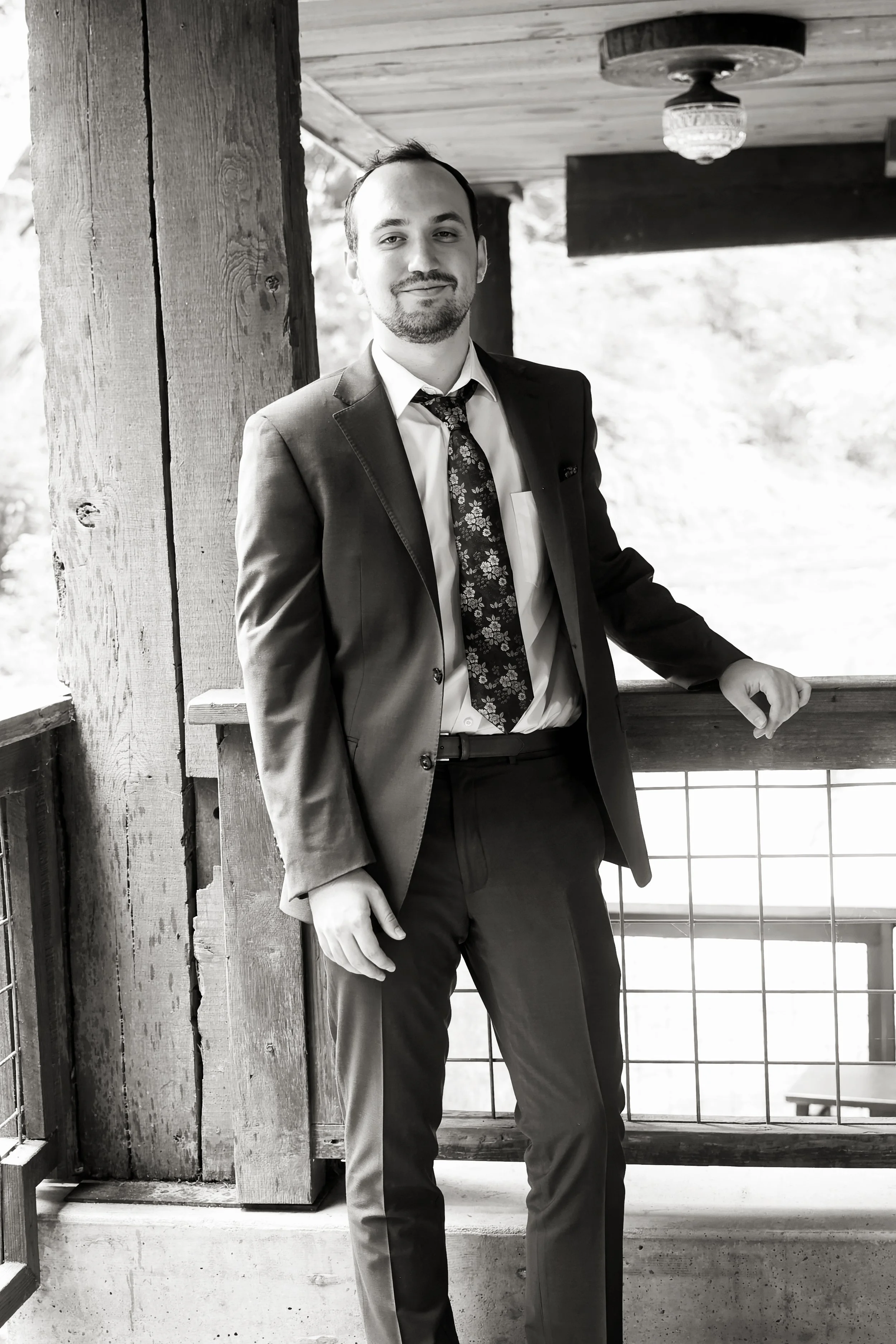 A young man in a suit and tie standing on a balcony with wooden beams, metal railing, and large grid windows in black and white.