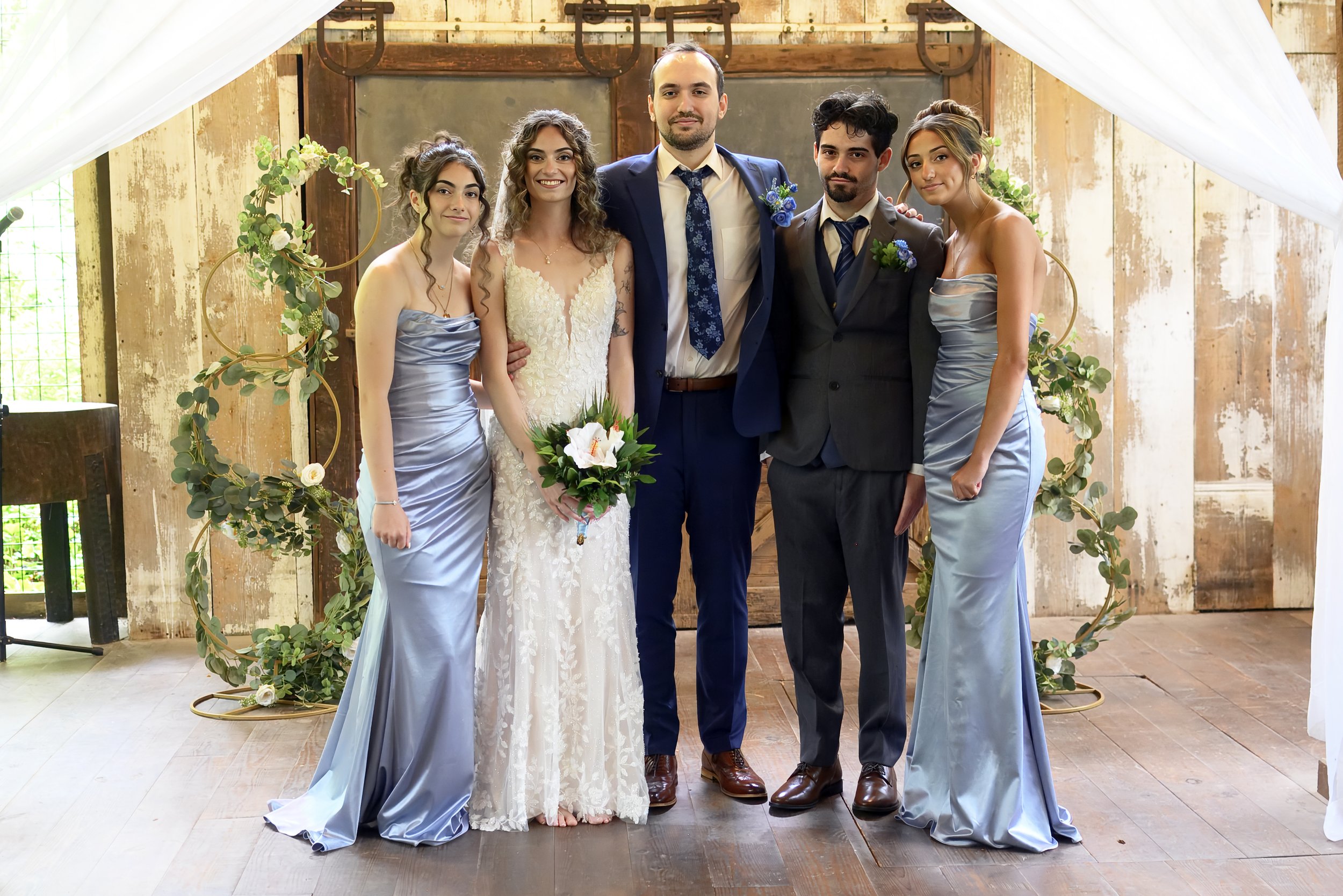 Group of five people at a wedding, standing under a white canopy with greenery and floral decorations, inside a rustic wooden building.