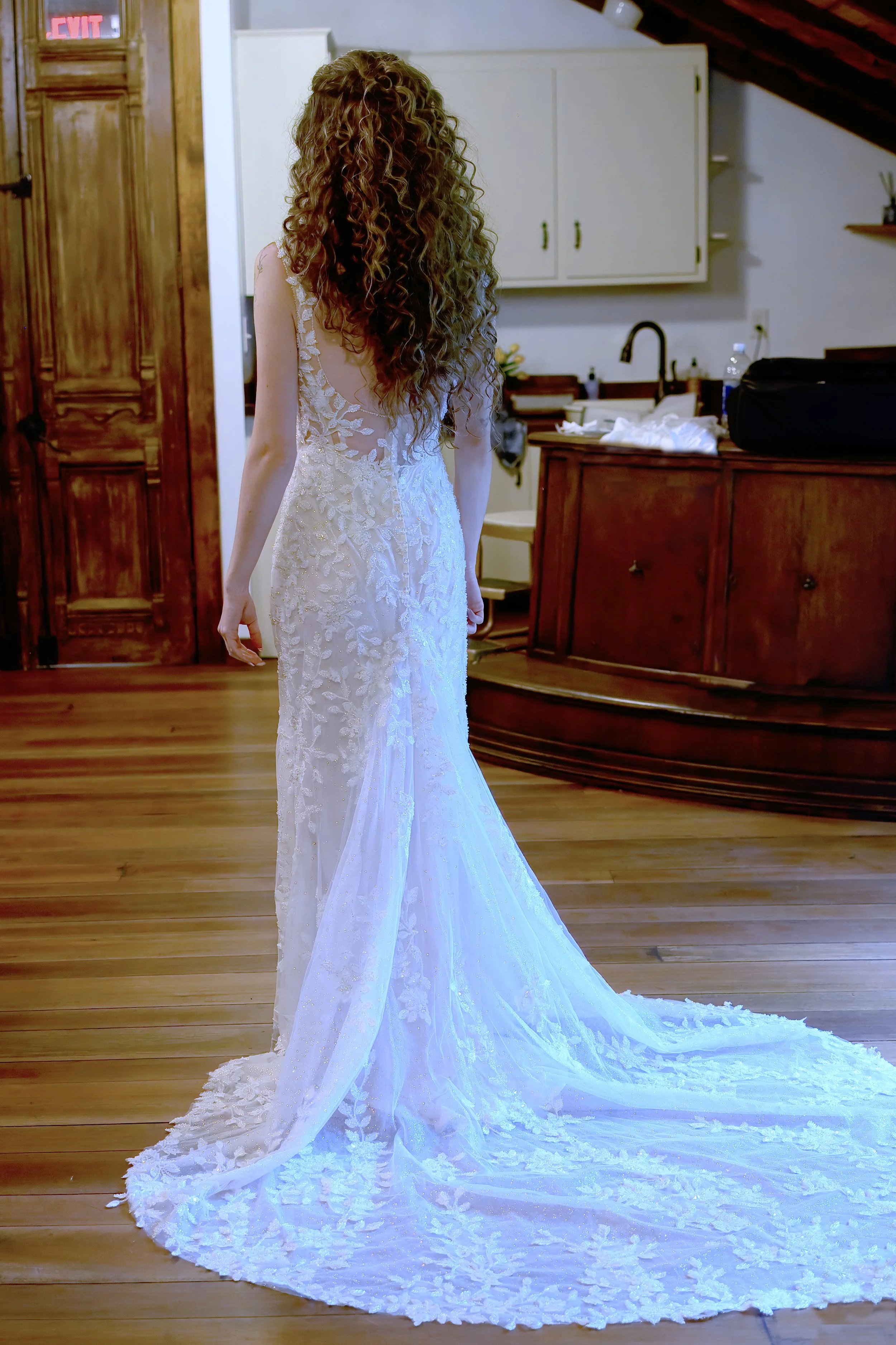 A woman with curly hair wearing a wedding dress standing indoors on wooden flooring, with a kitchen and wooden door in the background.