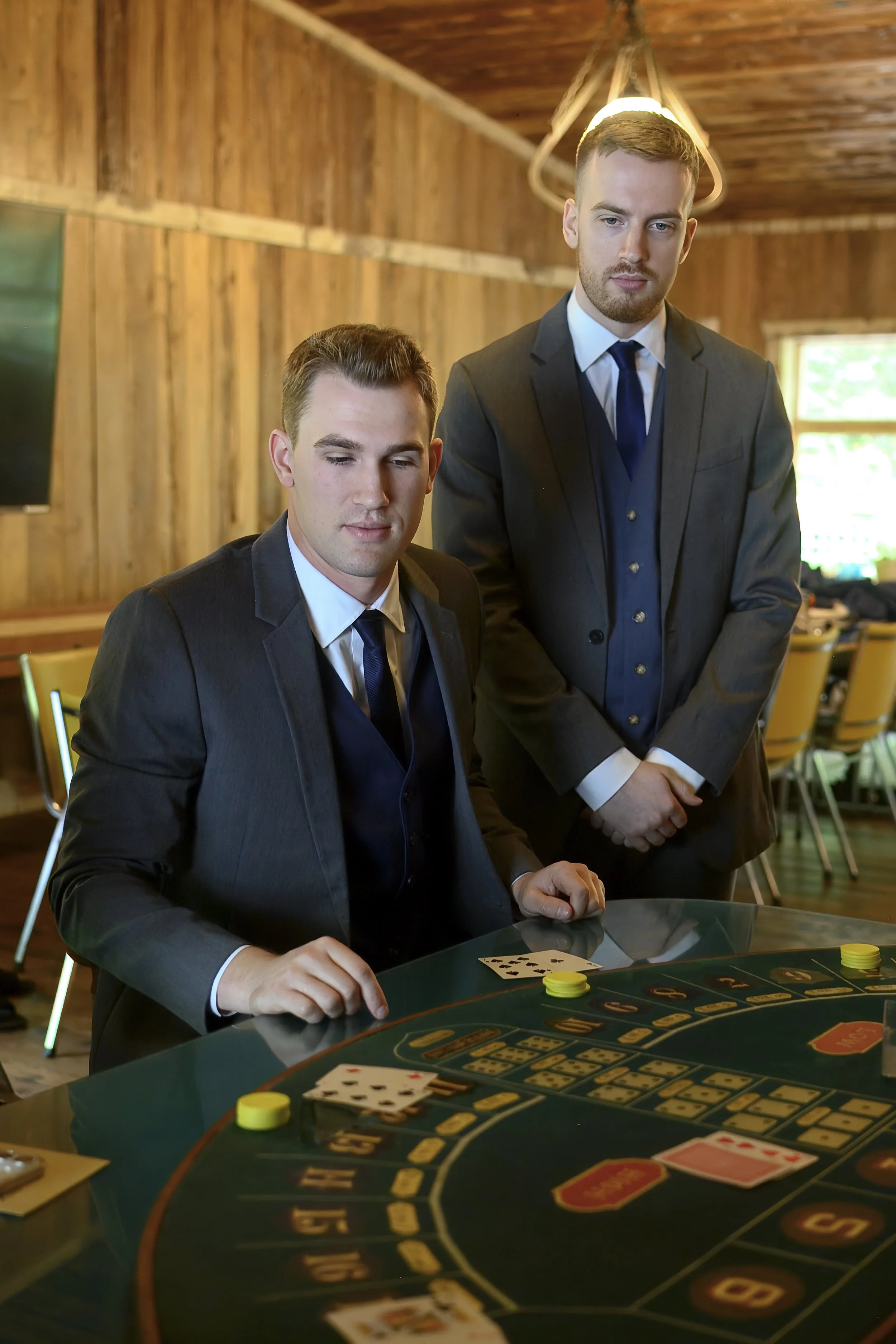 Two men in suits are playing blackjack at a casino table; one man is seated with playing cards and poker chips in front of him, while the other stands behind him, observing, in a cozy wood-paneled room with natural light.