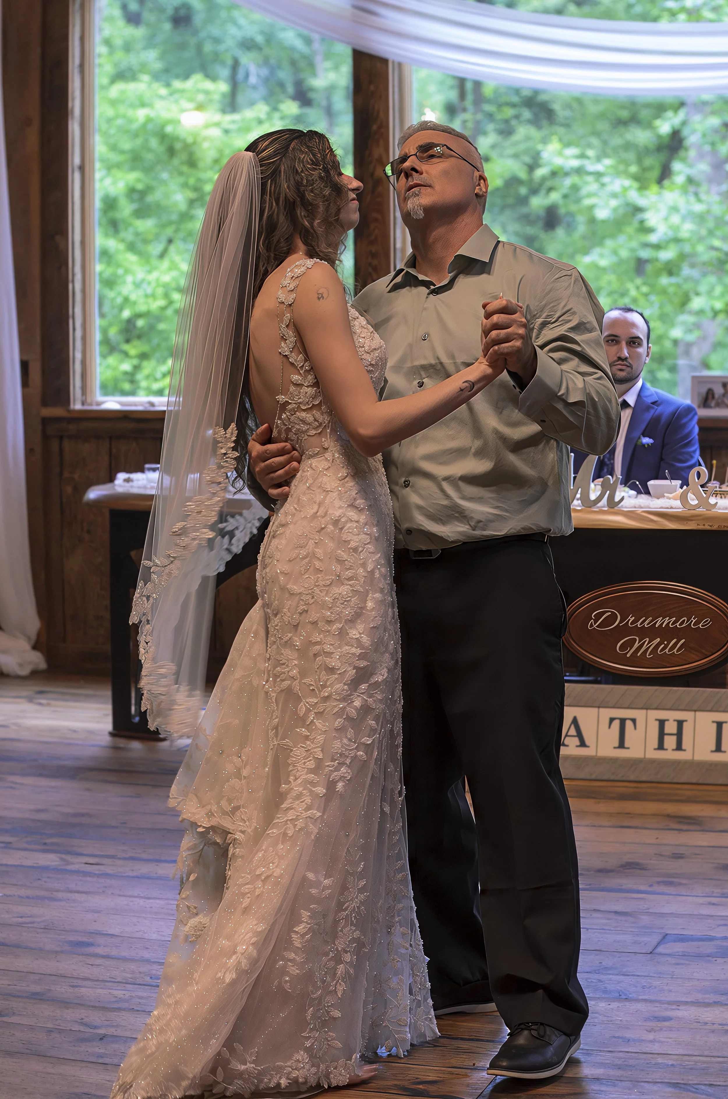 A bride and groom dancing at their wedding reception inside a rustic venue with large windows overlooking a green outdoor area. The bride is wearing a lace wedding gown with a veil, and the groom is dressed in a button-up shirt and dark pants. A man 