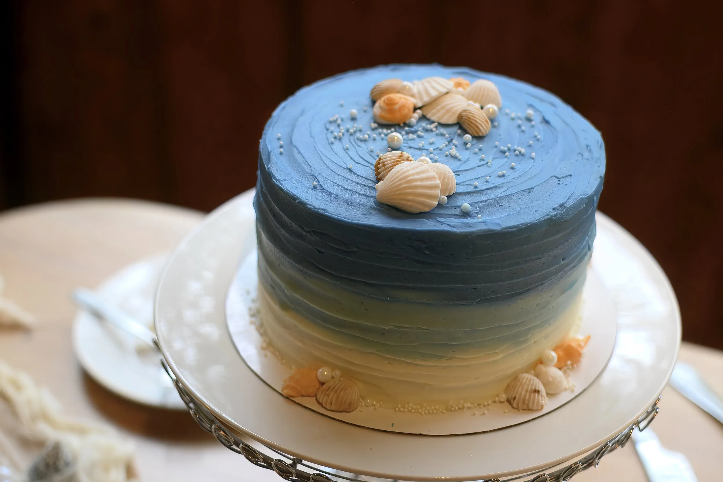 A multi-layered cake with blue gradient frosting decorated with seashell-shaped fondant and pearl-like edible decorations on top.