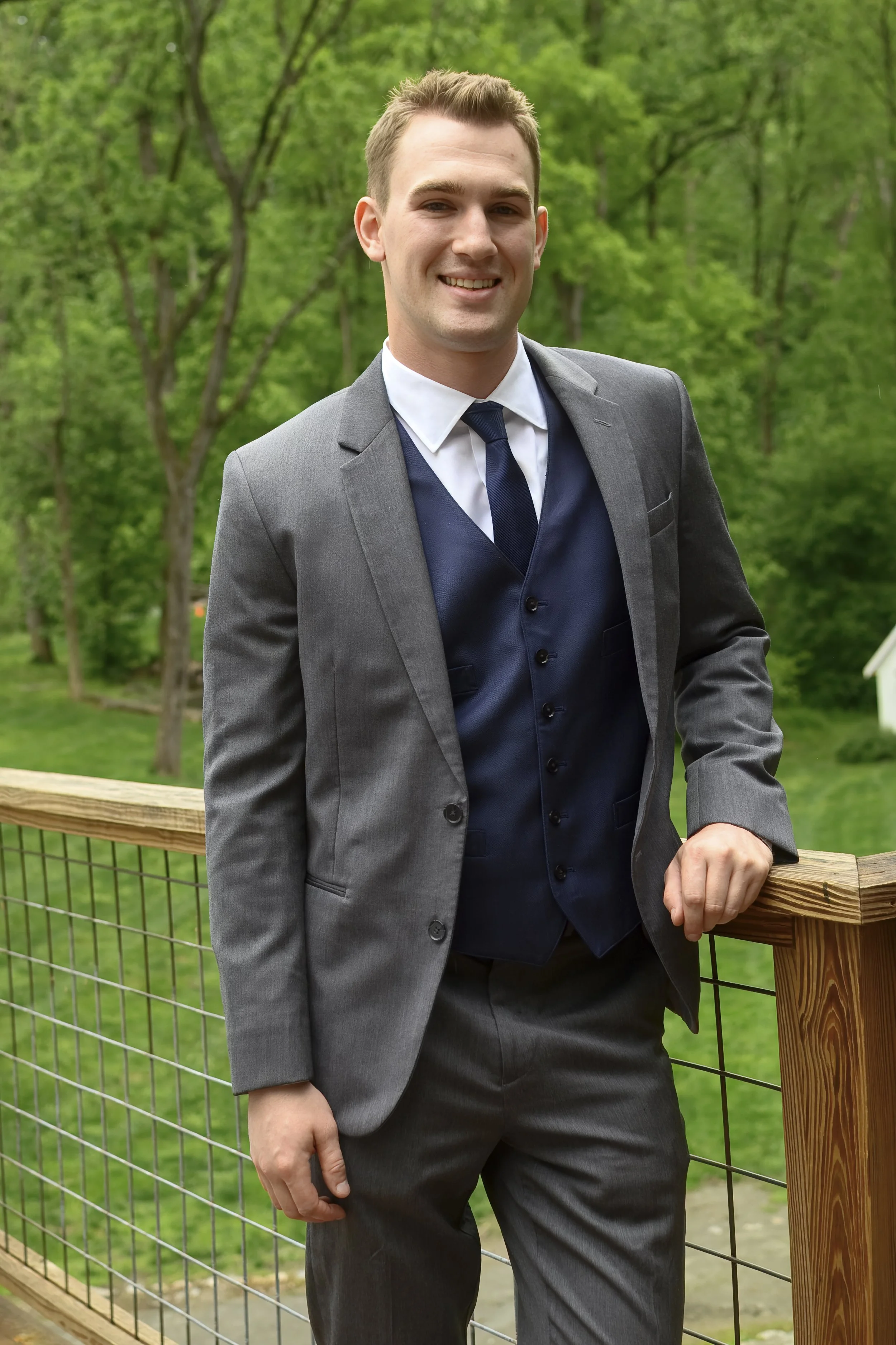 A young man in a gray suit with a dark blue vest and tie, smiling and standing outdoors by a wooden railing, with green trees in the background.