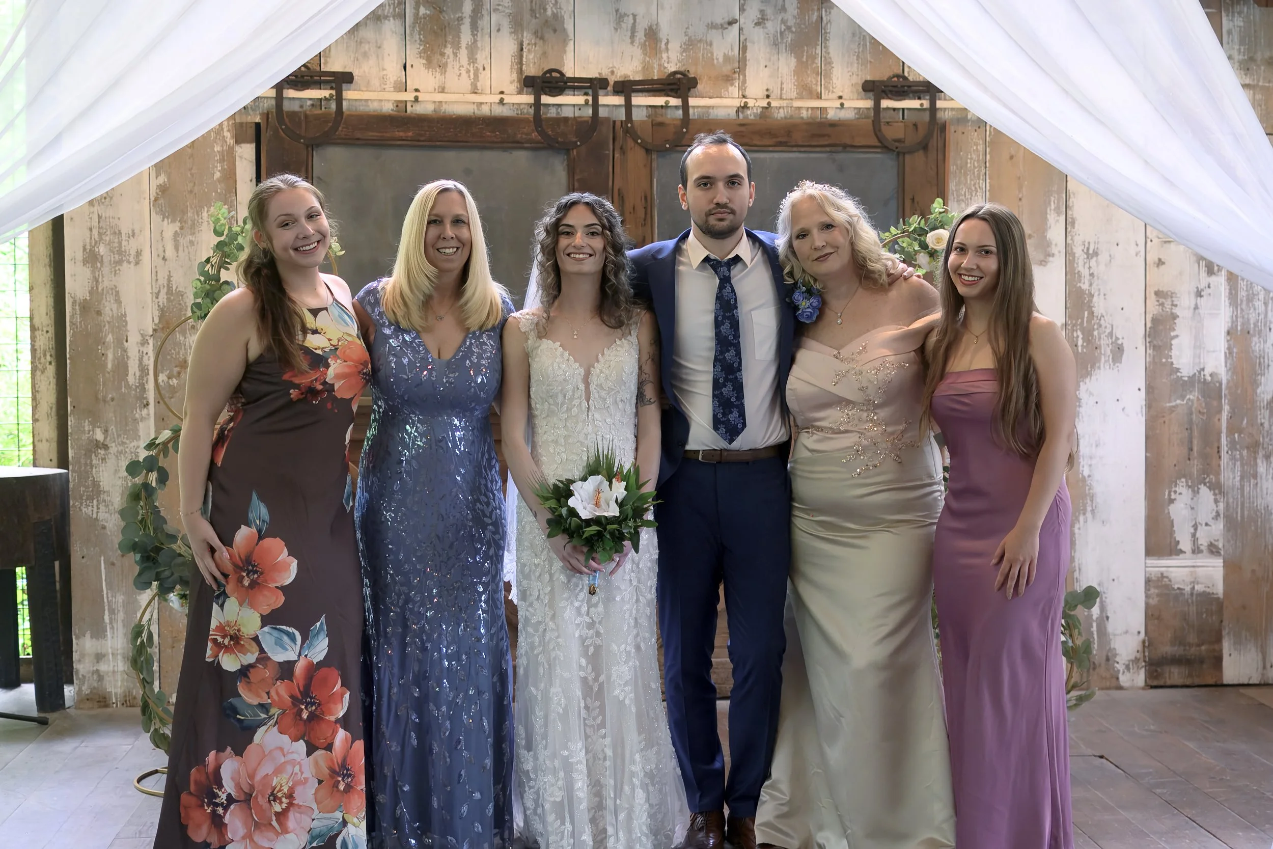 A group of six people, including a bride in a white wedding dress holding a bouquet, and five women in colorful dresses, standing together at a rustic wedding venue with wooden walls and greenery.