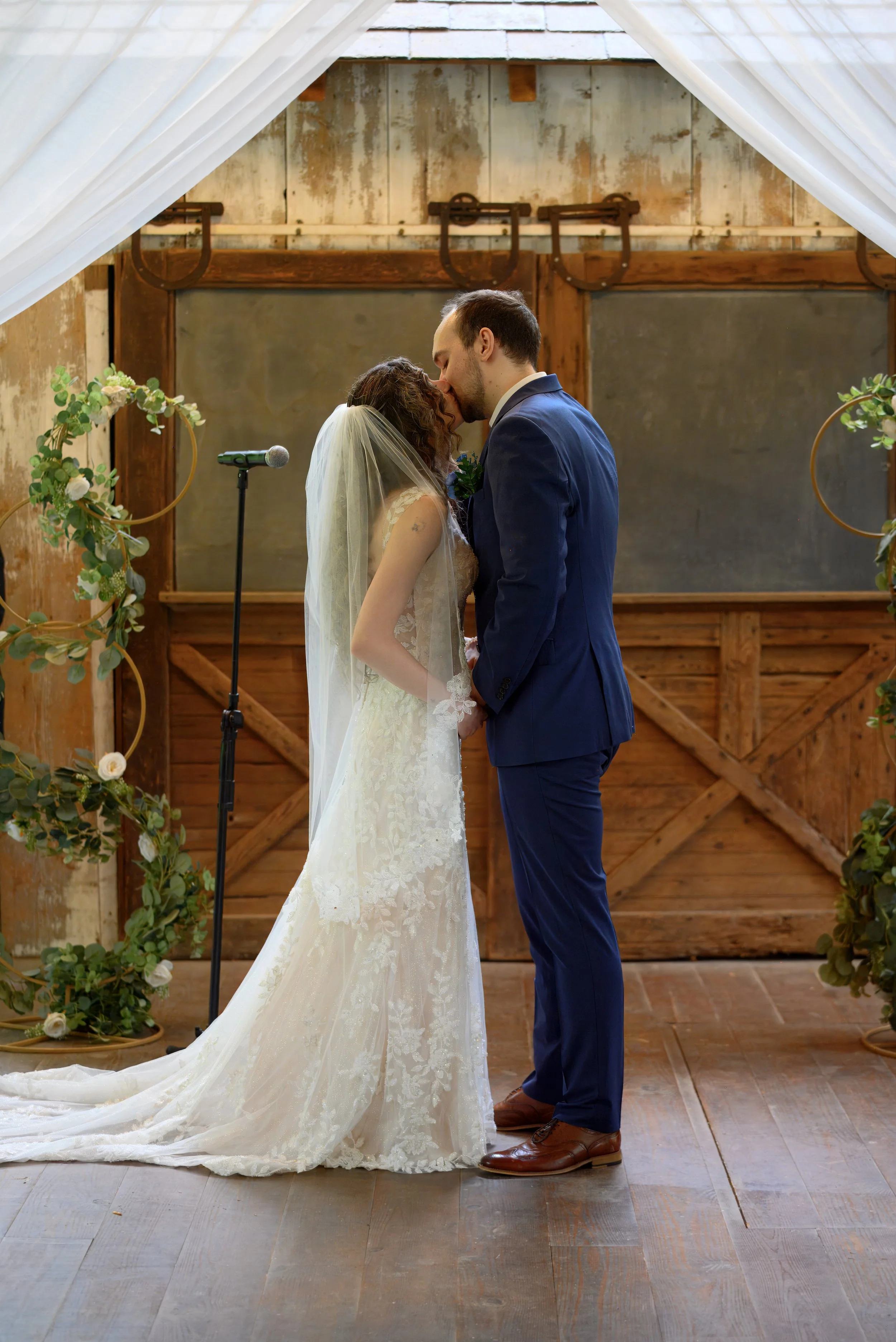 A bride and groom are sharing a kiss under a wedding arch decorated with greenery and white flowers in a rustic barn setting, with wooden walls and a large barn door in the background.