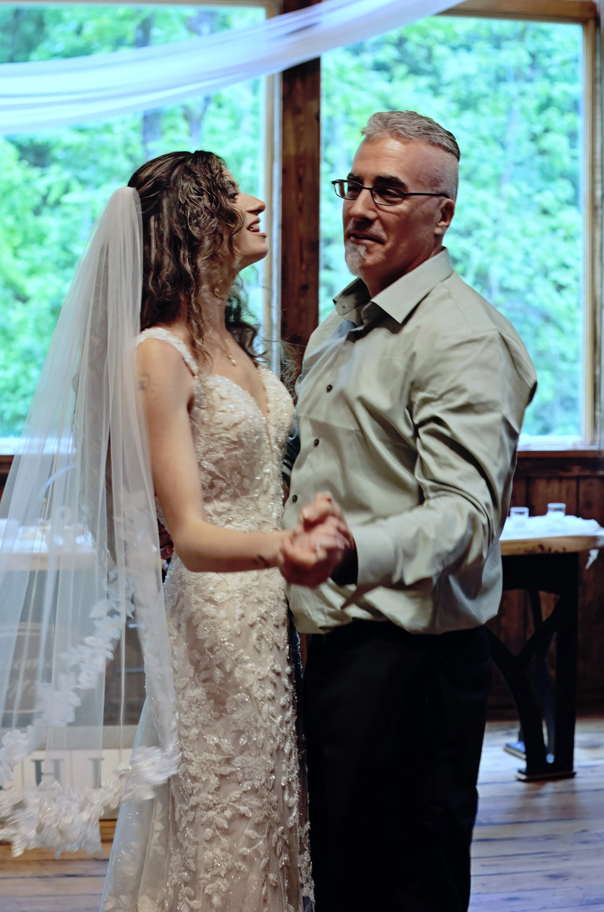 A bride and a man dancing together at a wedding reception, with a background of trees visible through large windows.