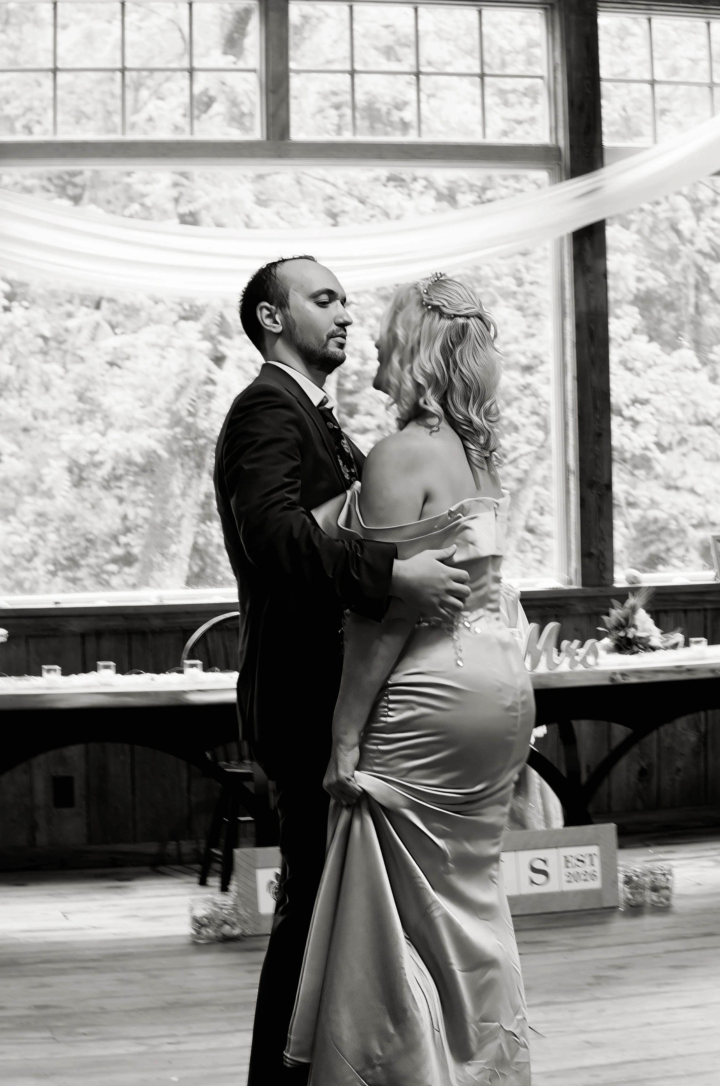 A couple, dressed in wedding attire, dancing closely indoors near large windows with a snowy landscape outside.