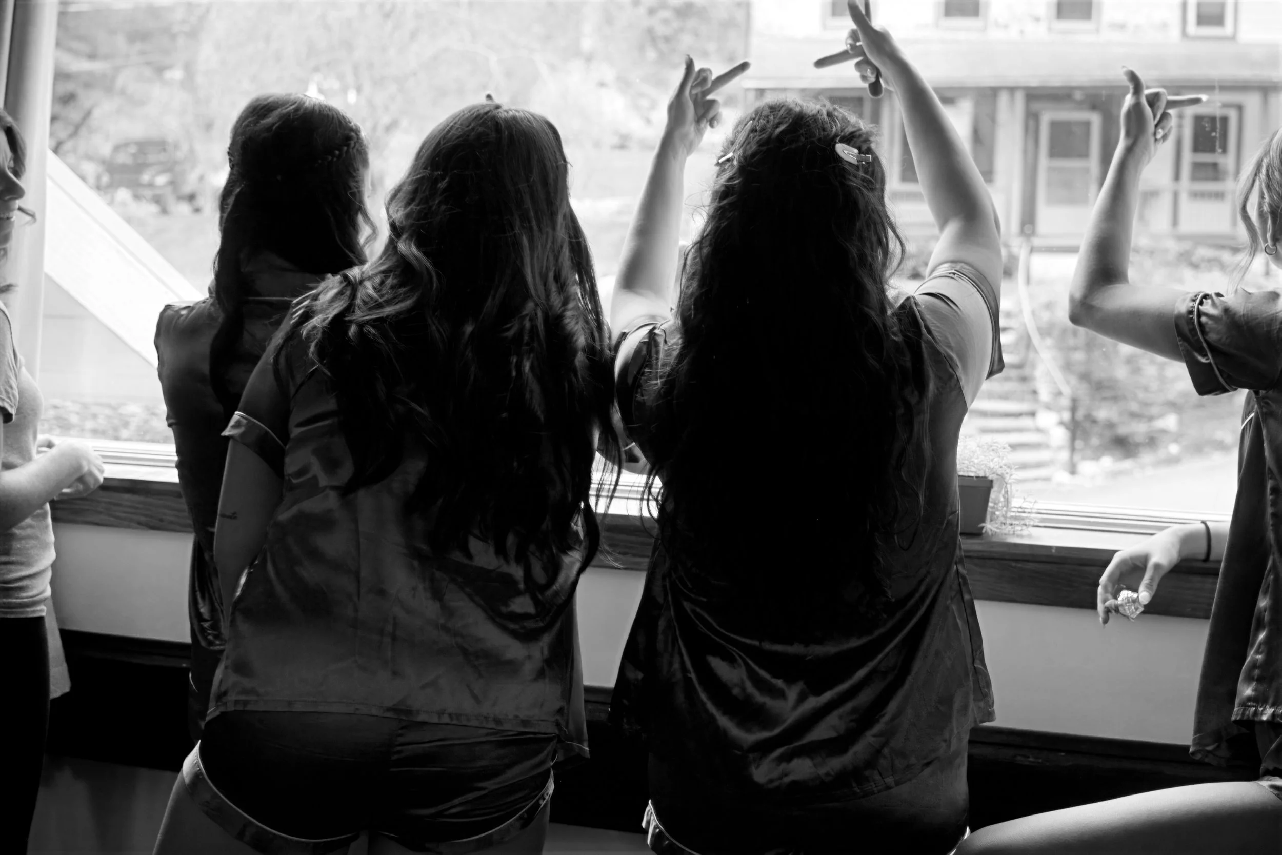 Group of women standing near a window, engaging in conversation and gestures, with a view of a residential street outside.