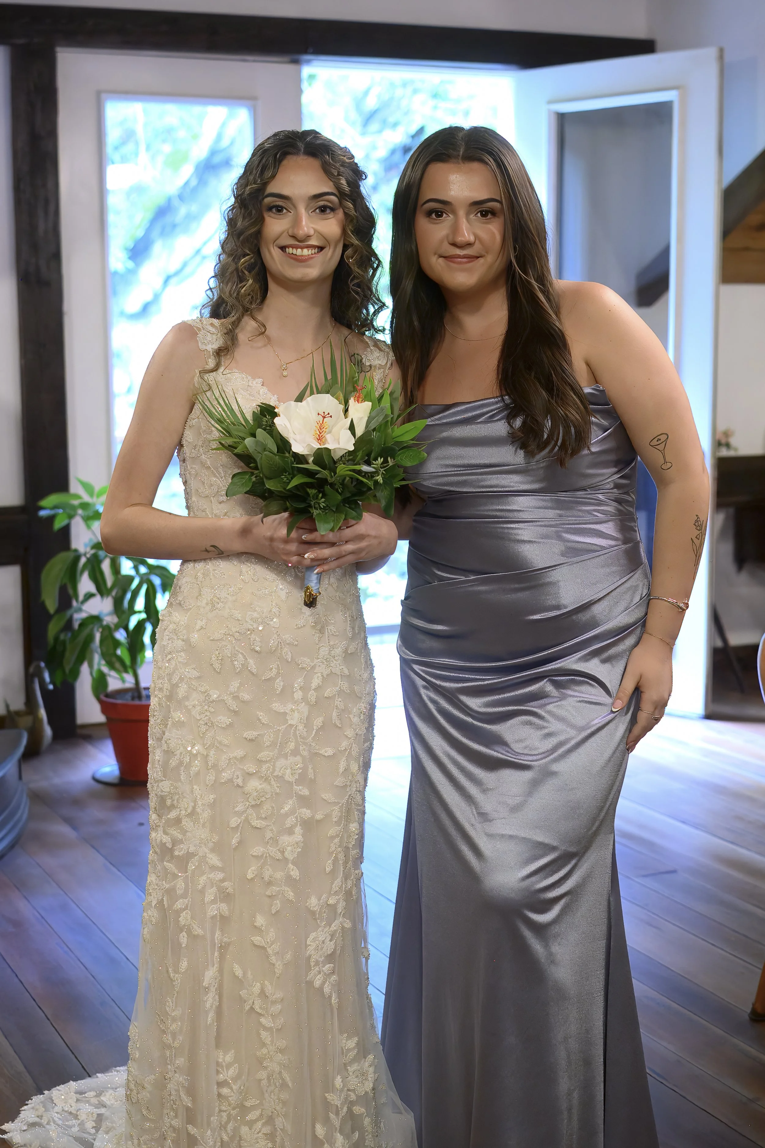 Two women standing together indoors, one in a wedding dress holding a bouquet of white flowers with greenery, smiling at the camera, and the other in a silver sleeveless gown with tattoos on her arms, also smiling, with a modern interior background a