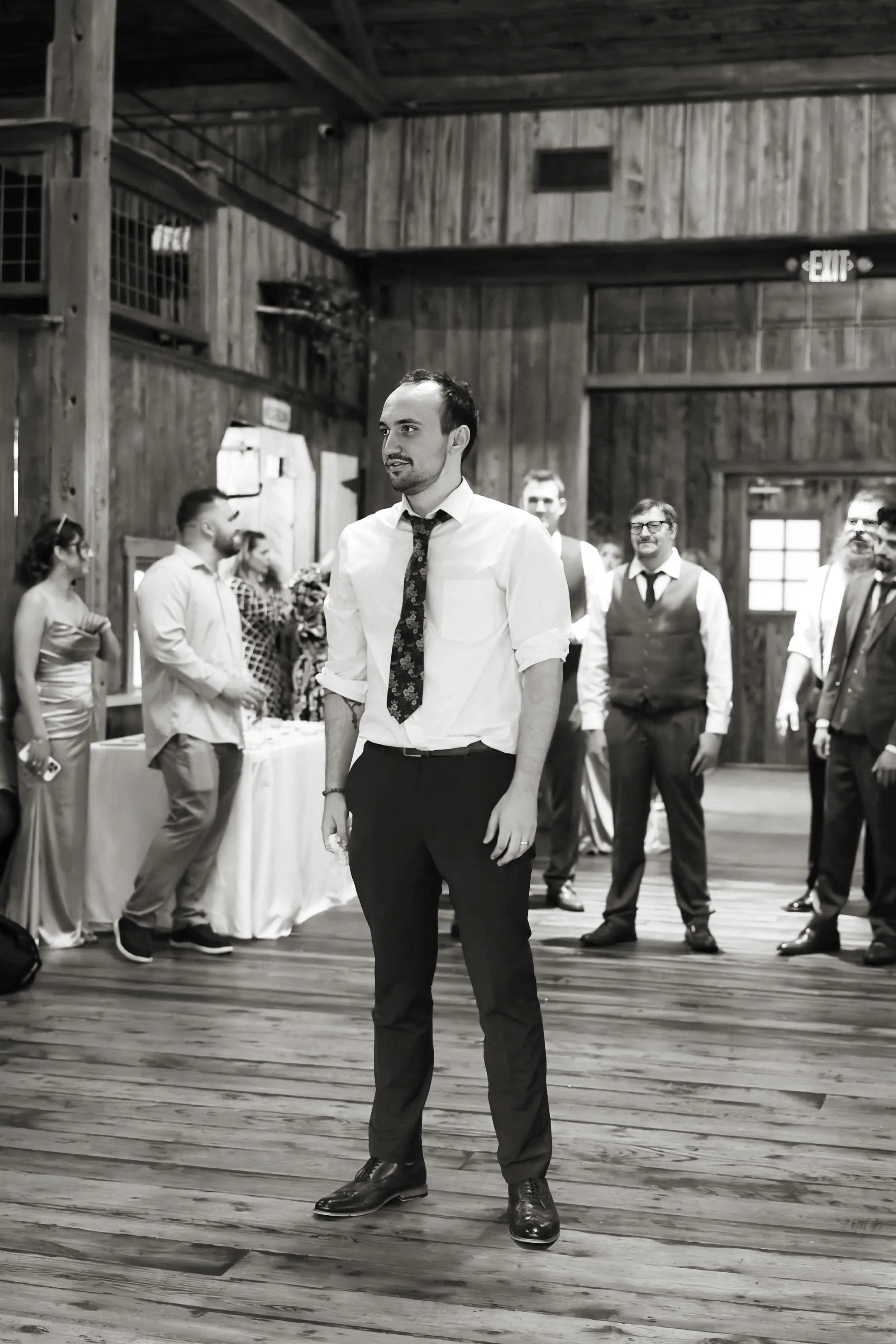 A man dressed in a shirt, tie, and dress pants standing in a wooden hall with surrounding people dressed in formal or semi-formal attire.