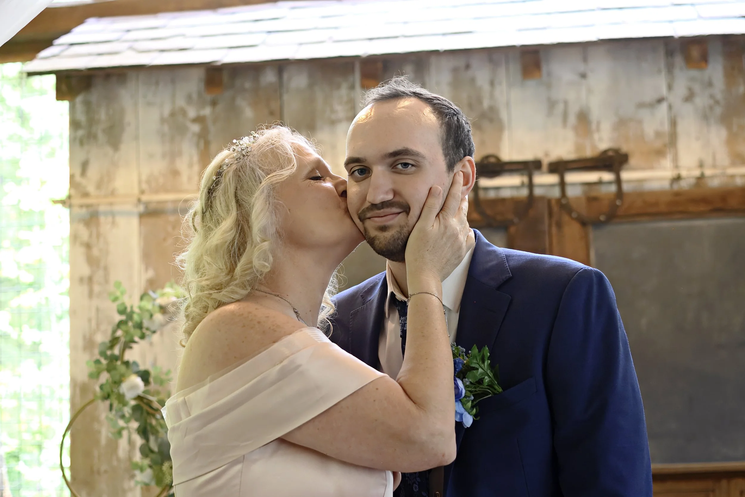 A woman with blonde curly hair kissing a young man on the cheek at a wedding. The woman is wearing a light-colored dress and a floral headband. The man is in a dark suit with a white shirt and tie. They are inside a rustic venue with wooden elements 