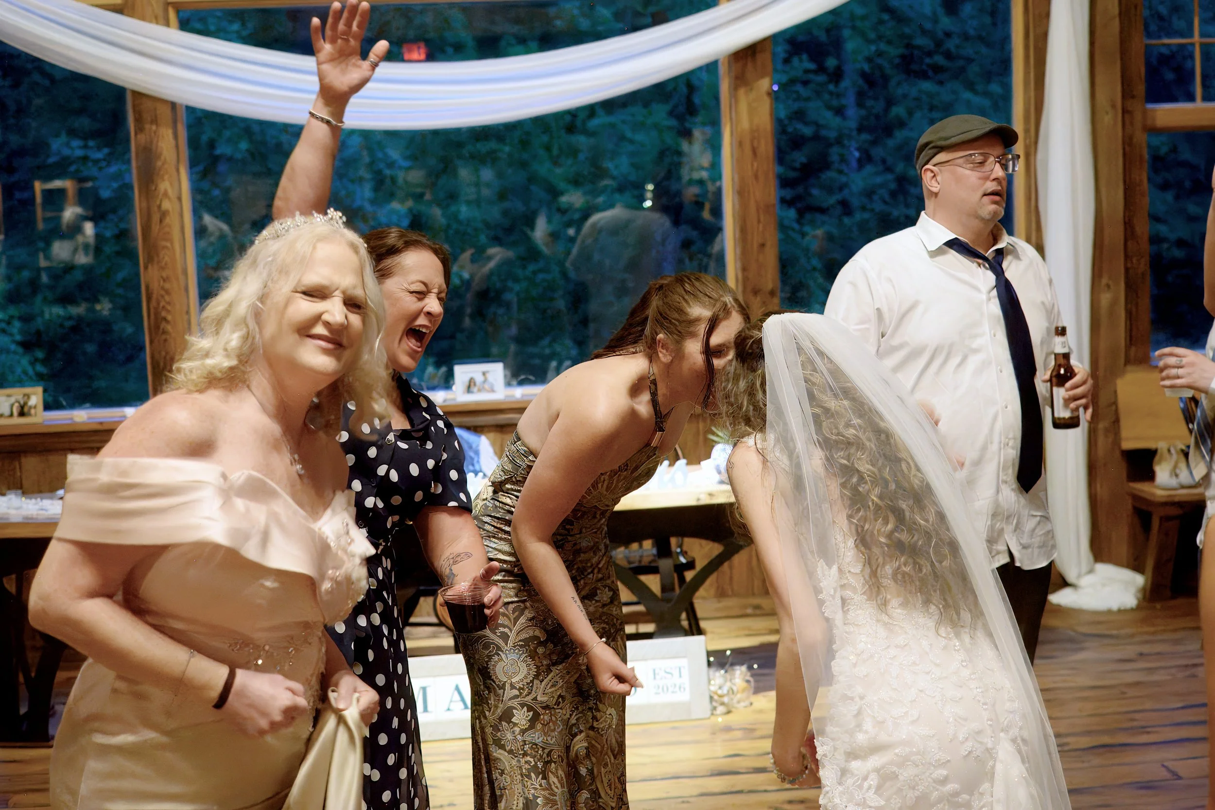 Group of people at an indoor celebration, some raising their hands, with a woman wearing a wedding dress and veil in the center. The setting has wooden beams and a large window showing trees outside.