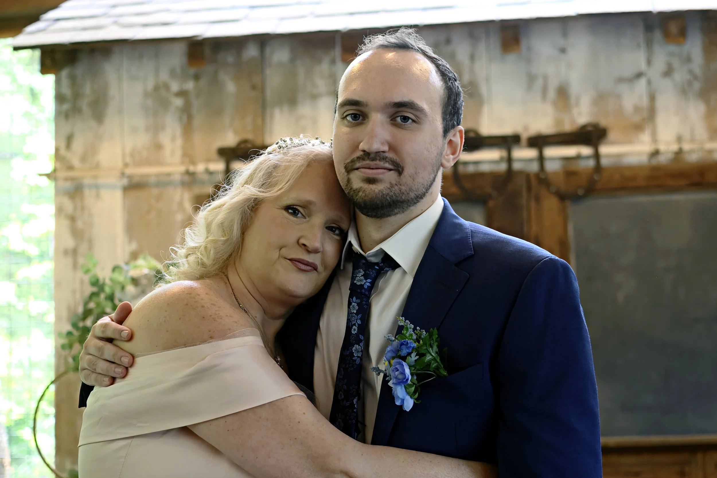 A man in a dark suit with a floral tie and boutonniere embraces a woman with blonde hair in a pink off-shoulder dress, both smiling, at a rustic indoor event with wooden beams and green foliage.