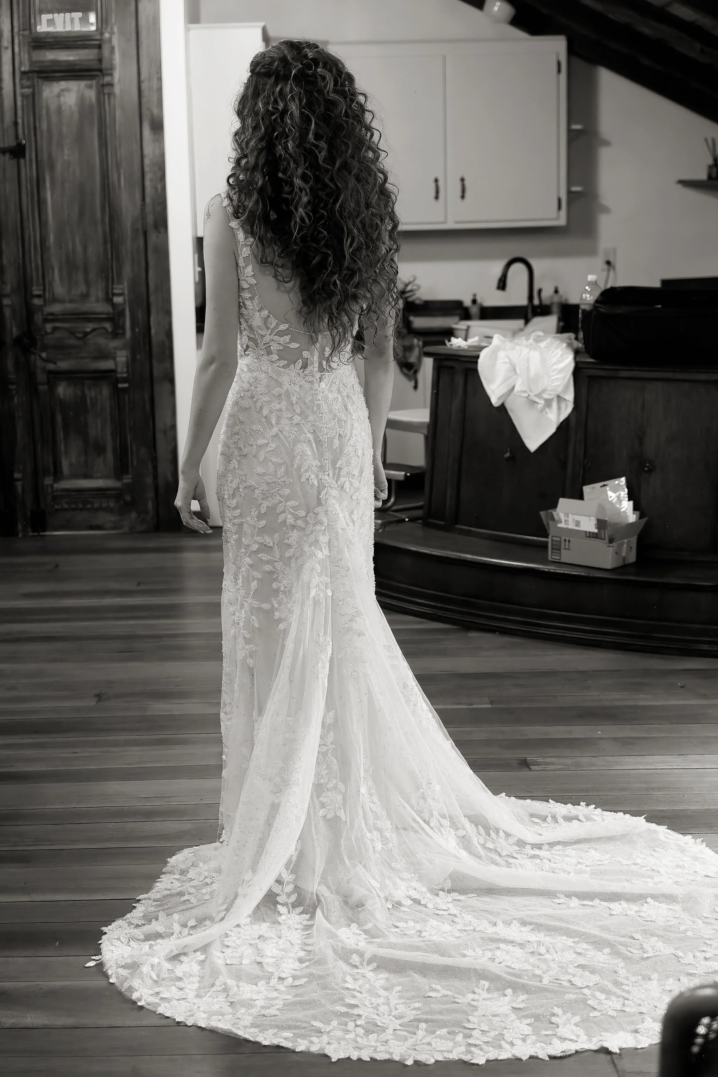 A woman in a wedding dress standing indoors with a long train, curly hair, and pull-up sleeves, in a room with wooden floors, white cabinets, and a cardboard box on the floor.