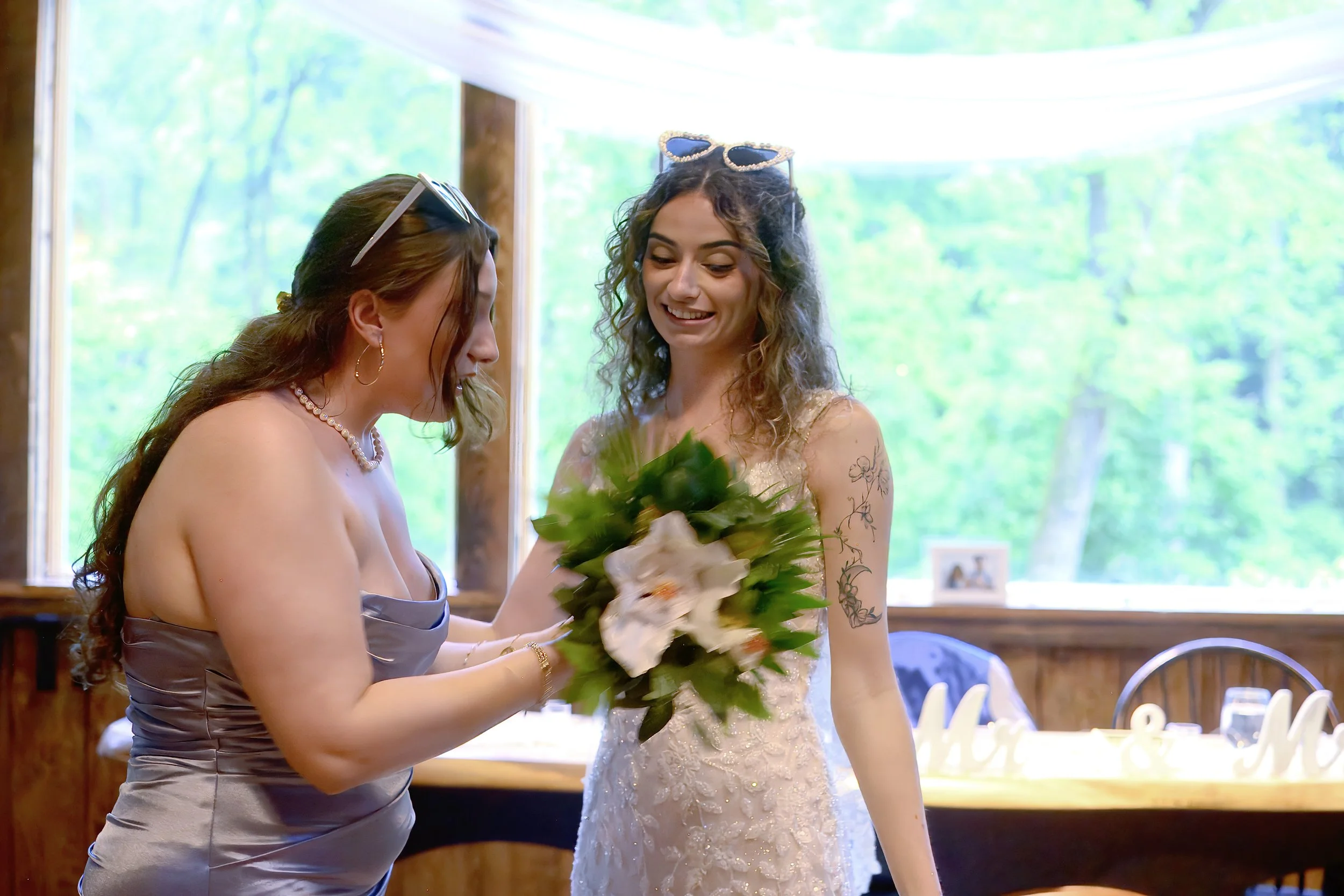 Two women smiling at each other, one holding a bouquet of flowers at a wedding reception with large windows and green trees outside.