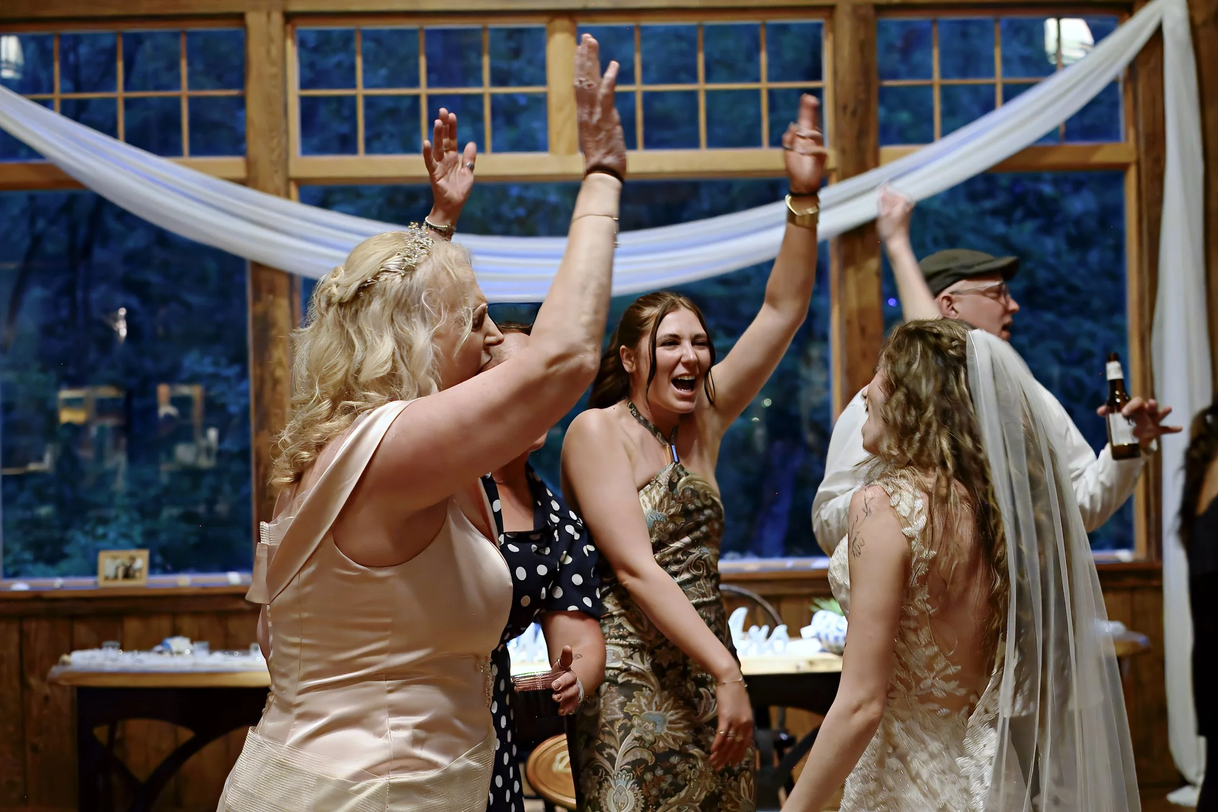 People celebrating at a wedding reception with their hands raised in a toast or cheer inside a wooden pavilion with large windows and white drapery.