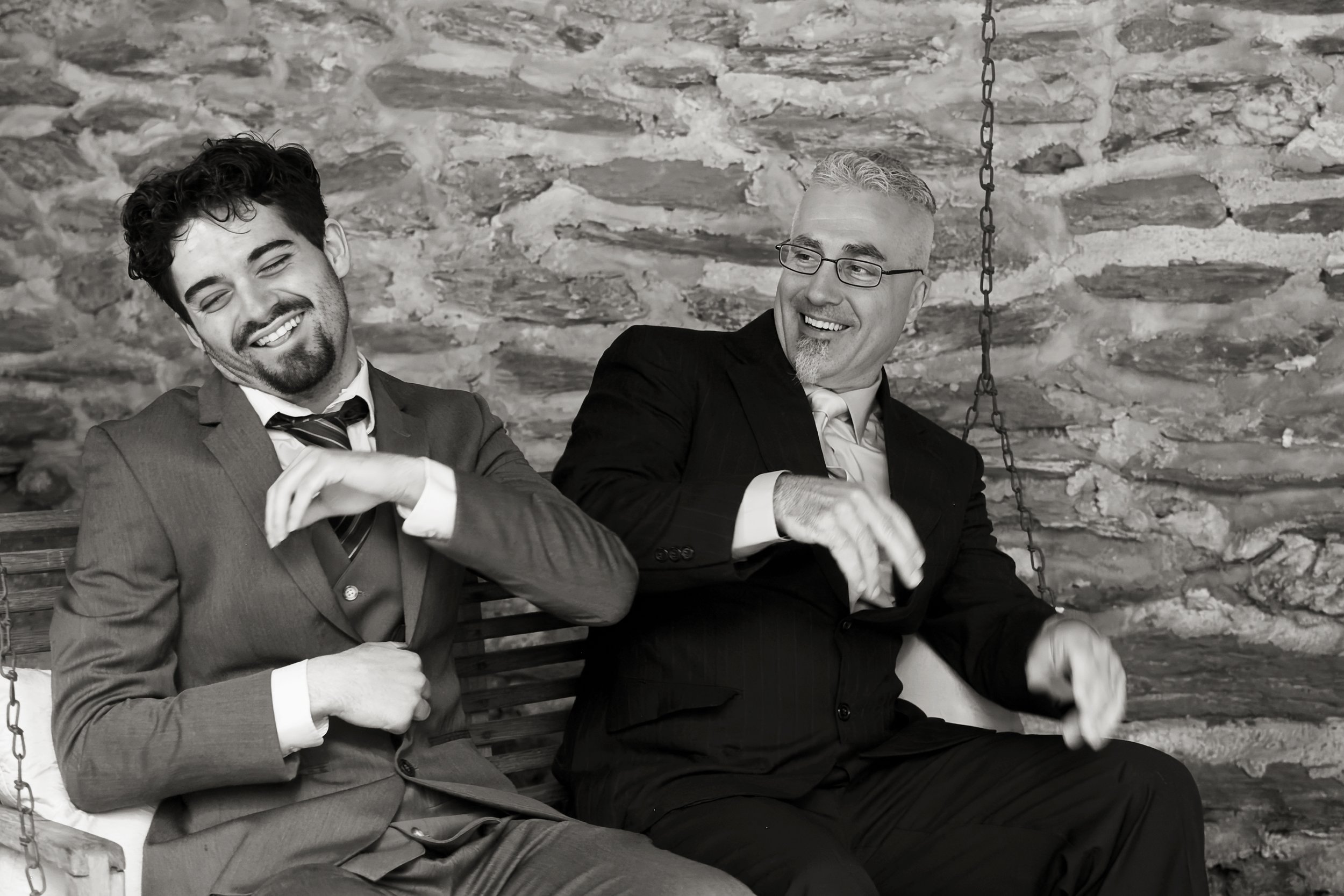 Two men in suits sitting on a swing with a stone wall background, smiling and talking.