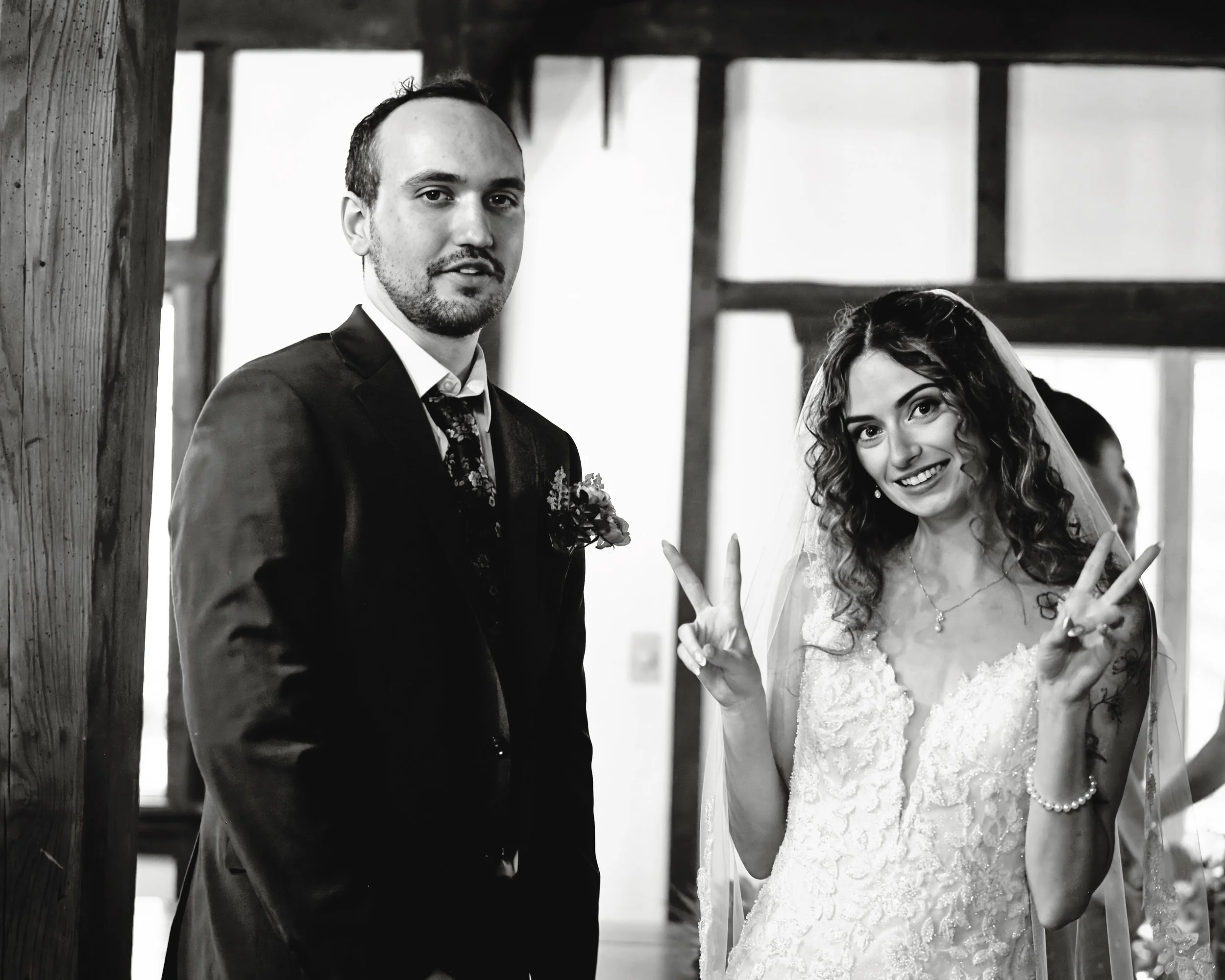 A black and white photo of a newlywed couple standing together inside a wooden structure, with the bride making a peace sign and smiling, and the groom standing beside her with a slight smile.