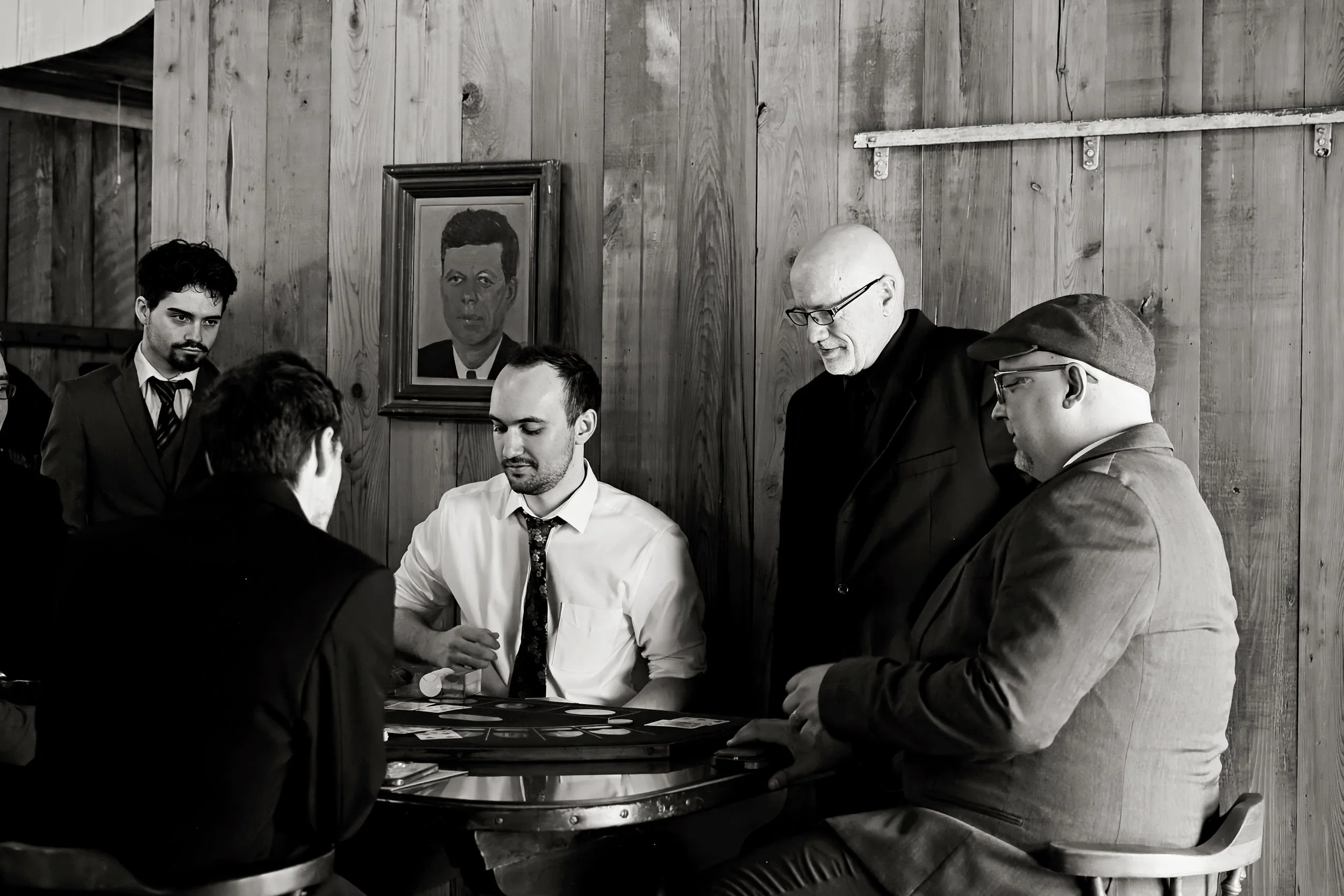 A group of men gathered around a table playing a card game in a room with wooden walls, with a framed portrait hanging on the wall behind them.