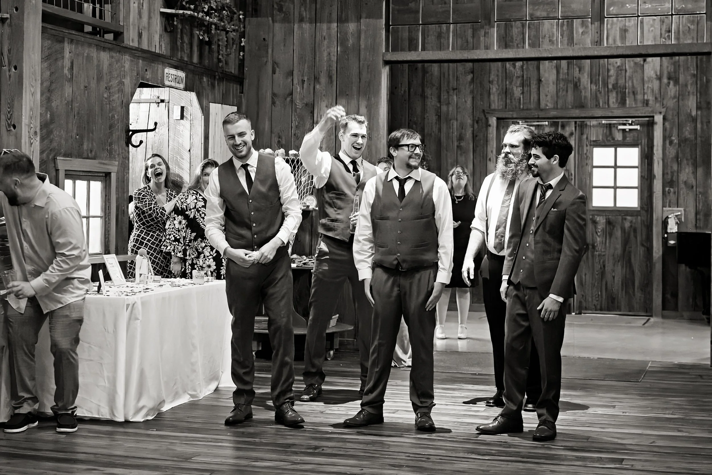 Group of smiling people in formal attire socializing at a rustic wooden indoor venue with tables and windows.
