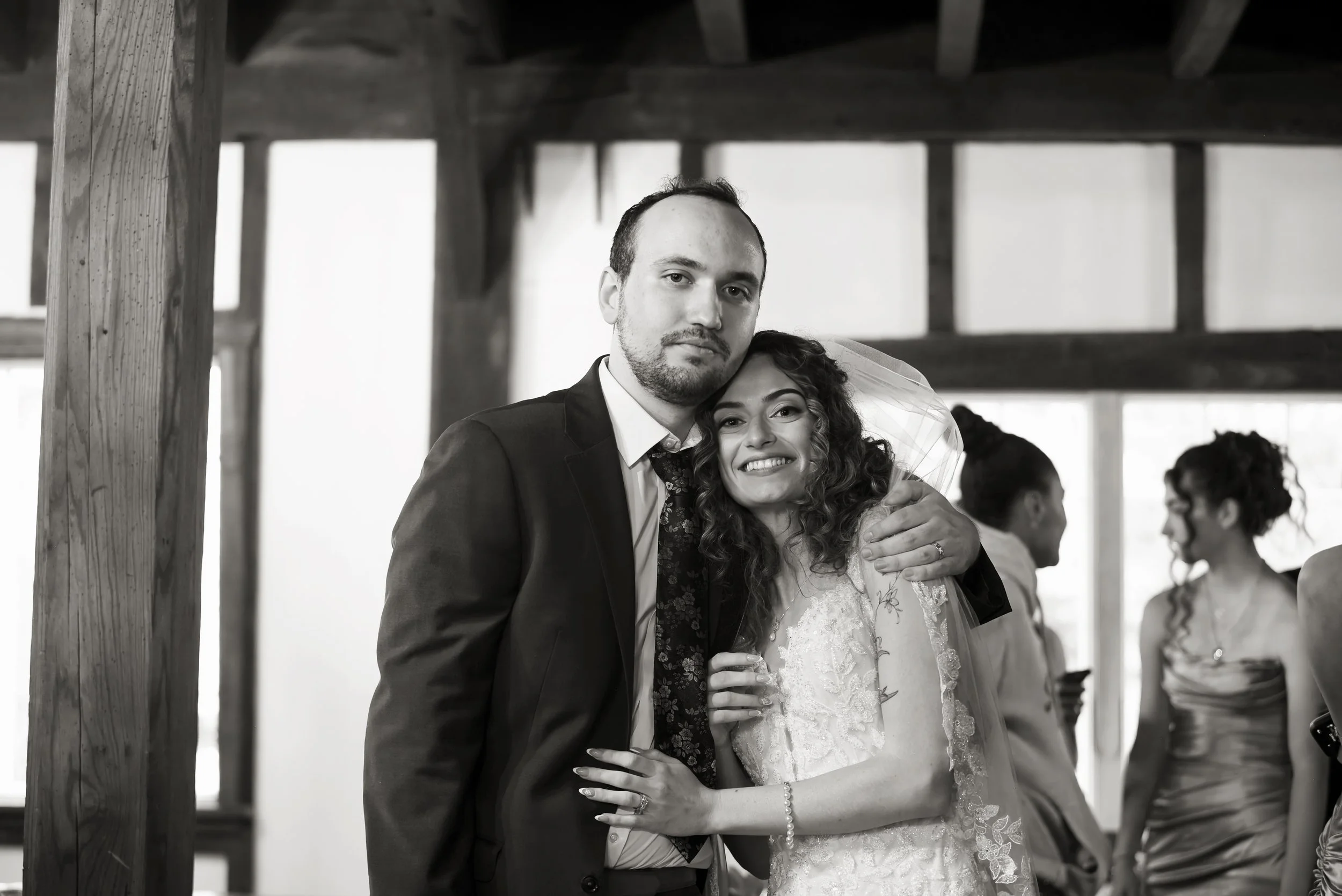 A couple, dressed in wedding attire, is hugging and smiling at an indoor wedding reception. The man is wearing a dark suit with a white shirt and floral tie, and the woman is wearing a lace wedding dress with a veil. There are other guests in the bac
