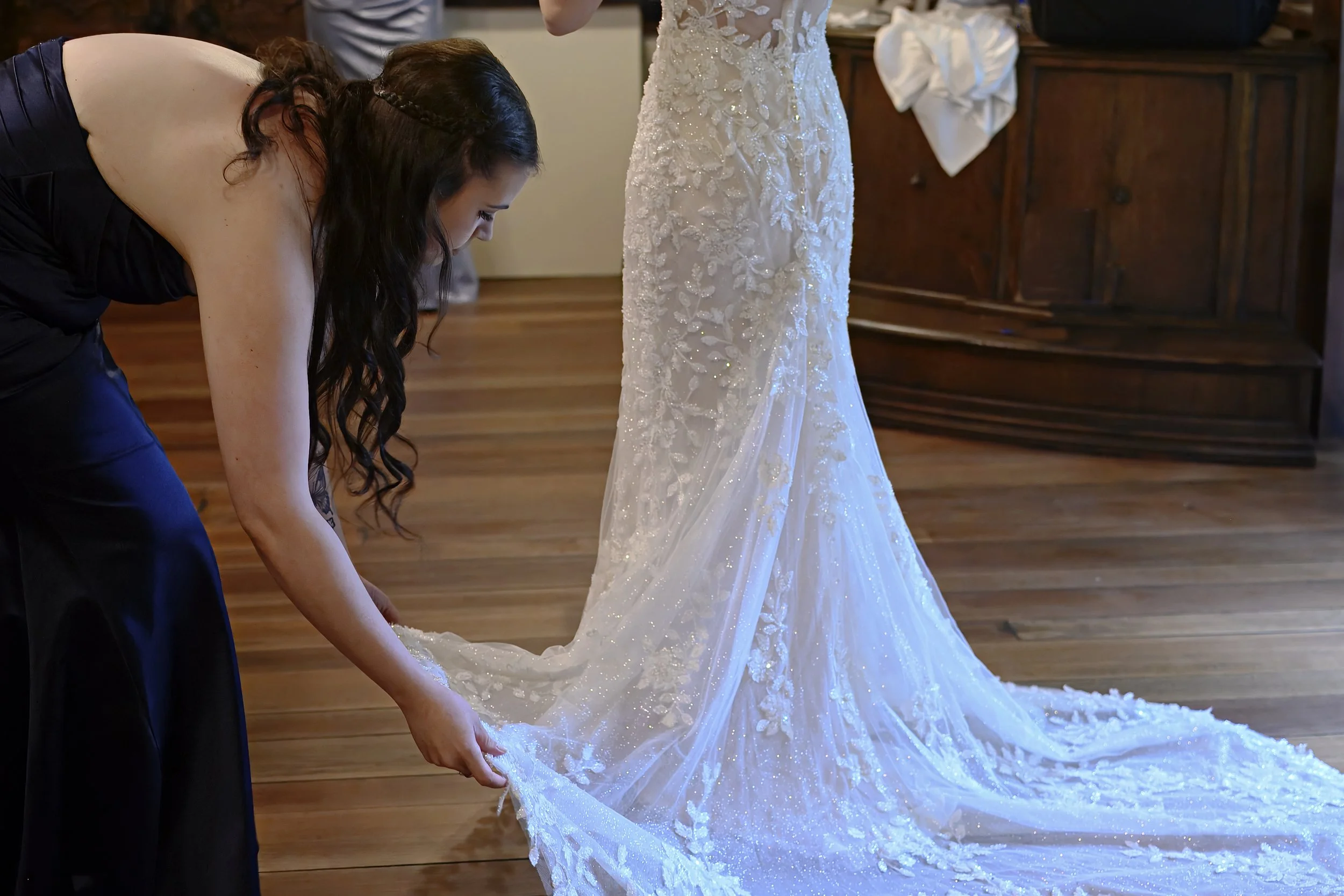 A woman in a black dress is bending down to adjust the train of her white, beaded wedding gown. The scene appears to be inside a wooden-floored room, possibly a wedding venue or dressing room.