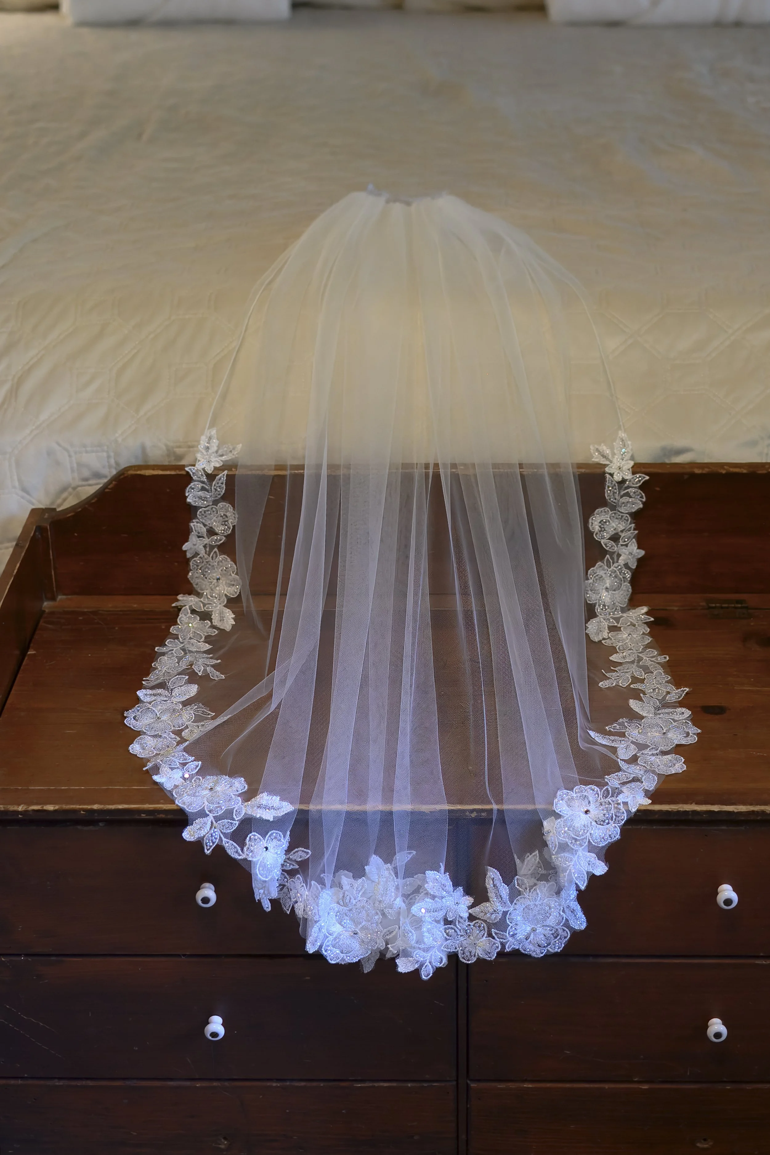 A white wedding veil with lace and floral embroidery, draped over a wooden dresser with small white knobs.