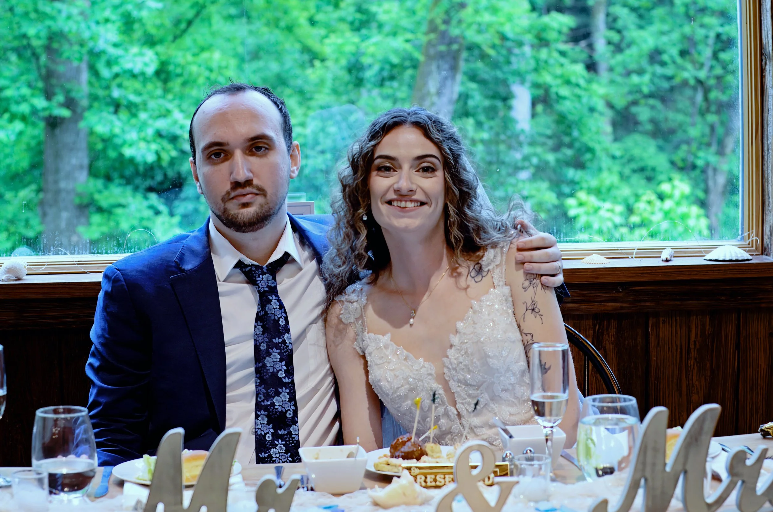 A smiling woman and a man sit closely together at a table during a celebration, with a scenic green forest visible through a large window behind them. The woman wears a white lace dress, and the man is dressed in a dark suit with a patterned tie.