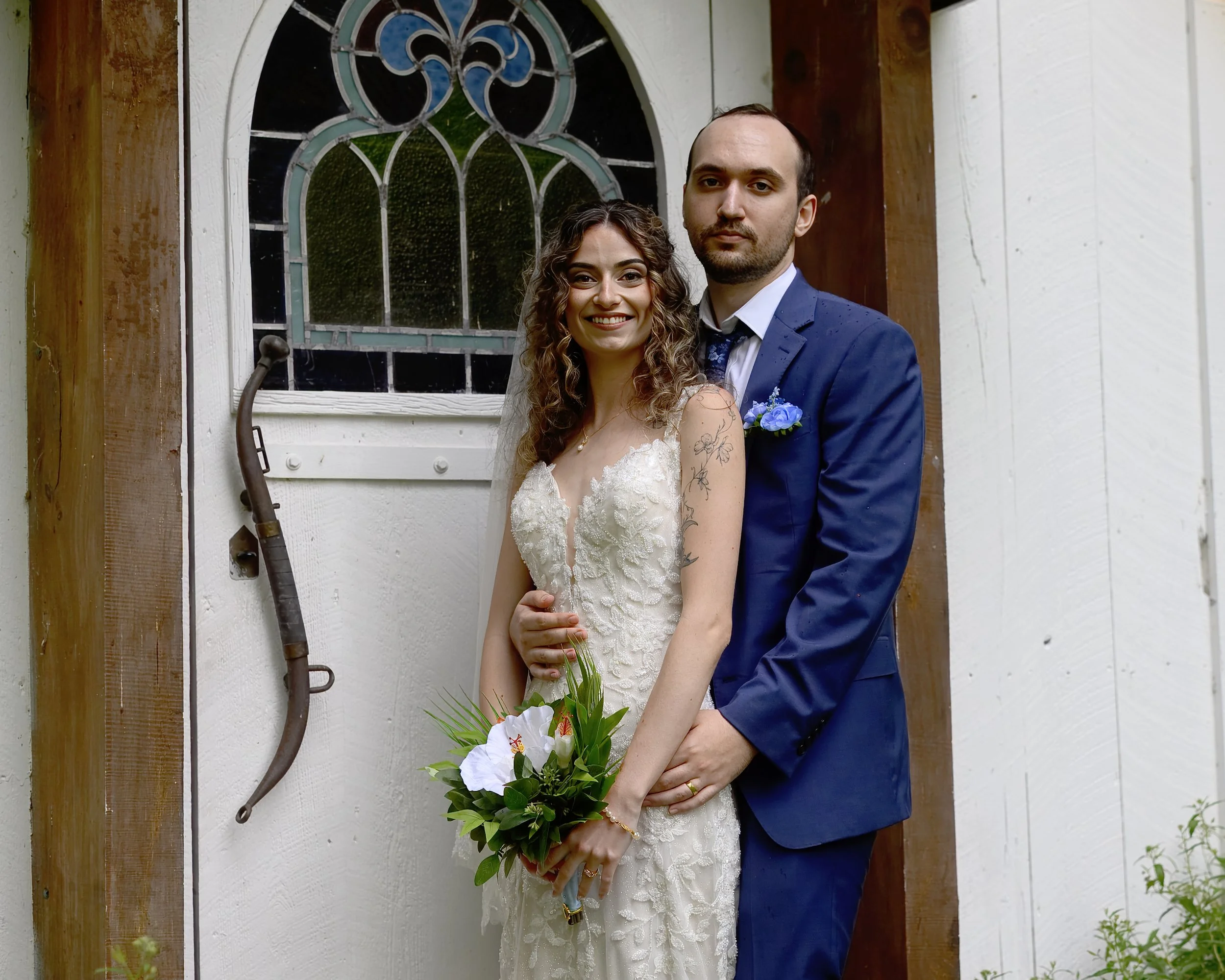 A bride and groom standing close together, smiling at the camera outside a house with a white wall and stained glass window. The bride has curly hair and is wearing a wedding dress holding a bouquet of white flowers with greenery. The groom has short