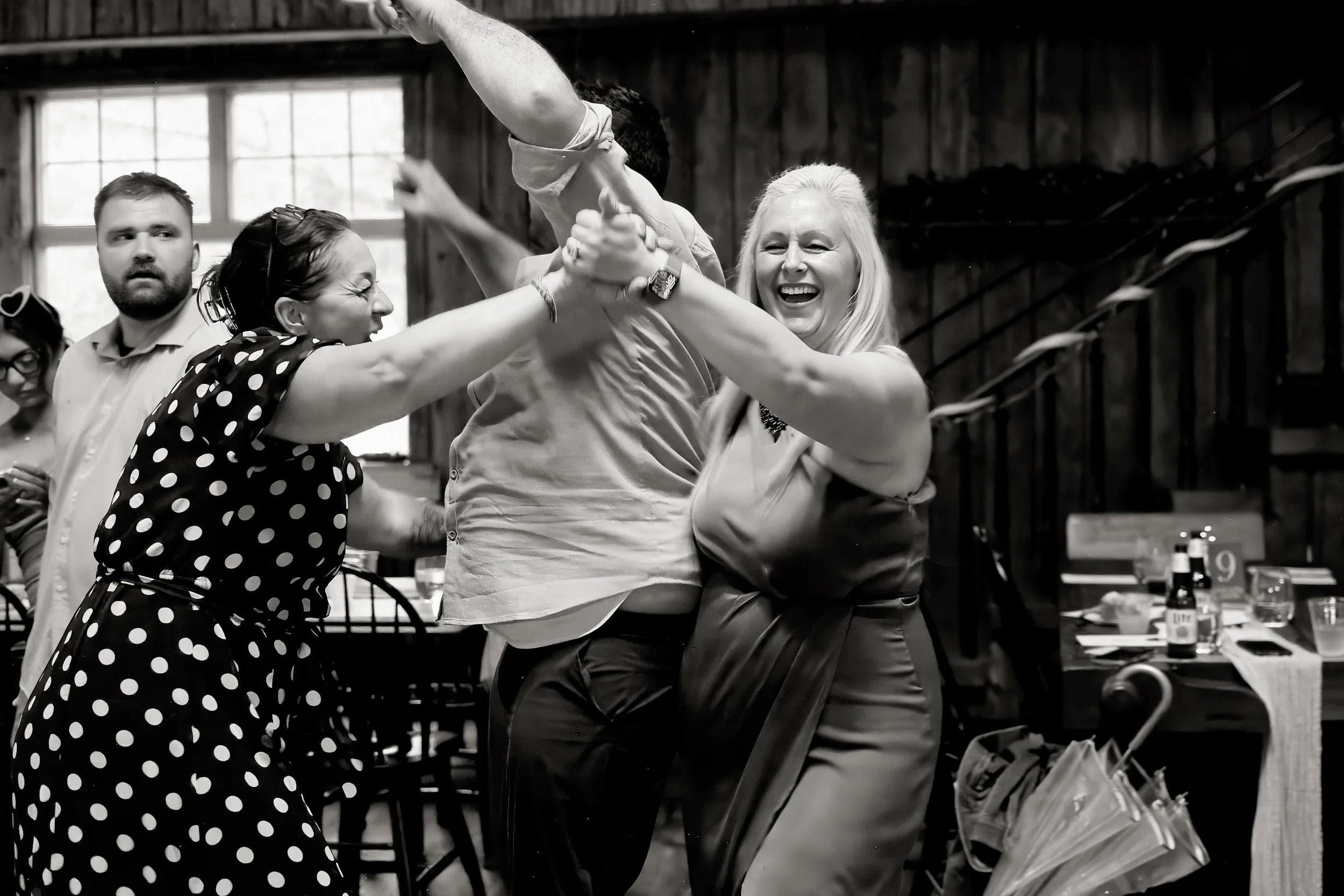 Two women are in a playful, physical fight during a celebration or party, with smiles and laughter, in a rustic indoor setting.
