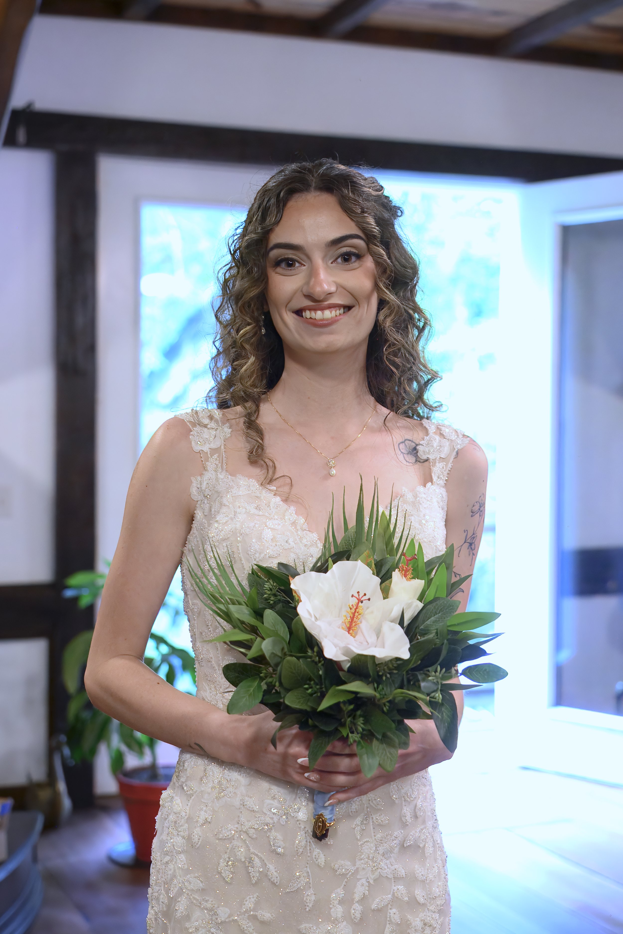 A woman in a wedding dress holding a bouquet of white flowers and green foliage, standing inside in front of a window with blue lighting.