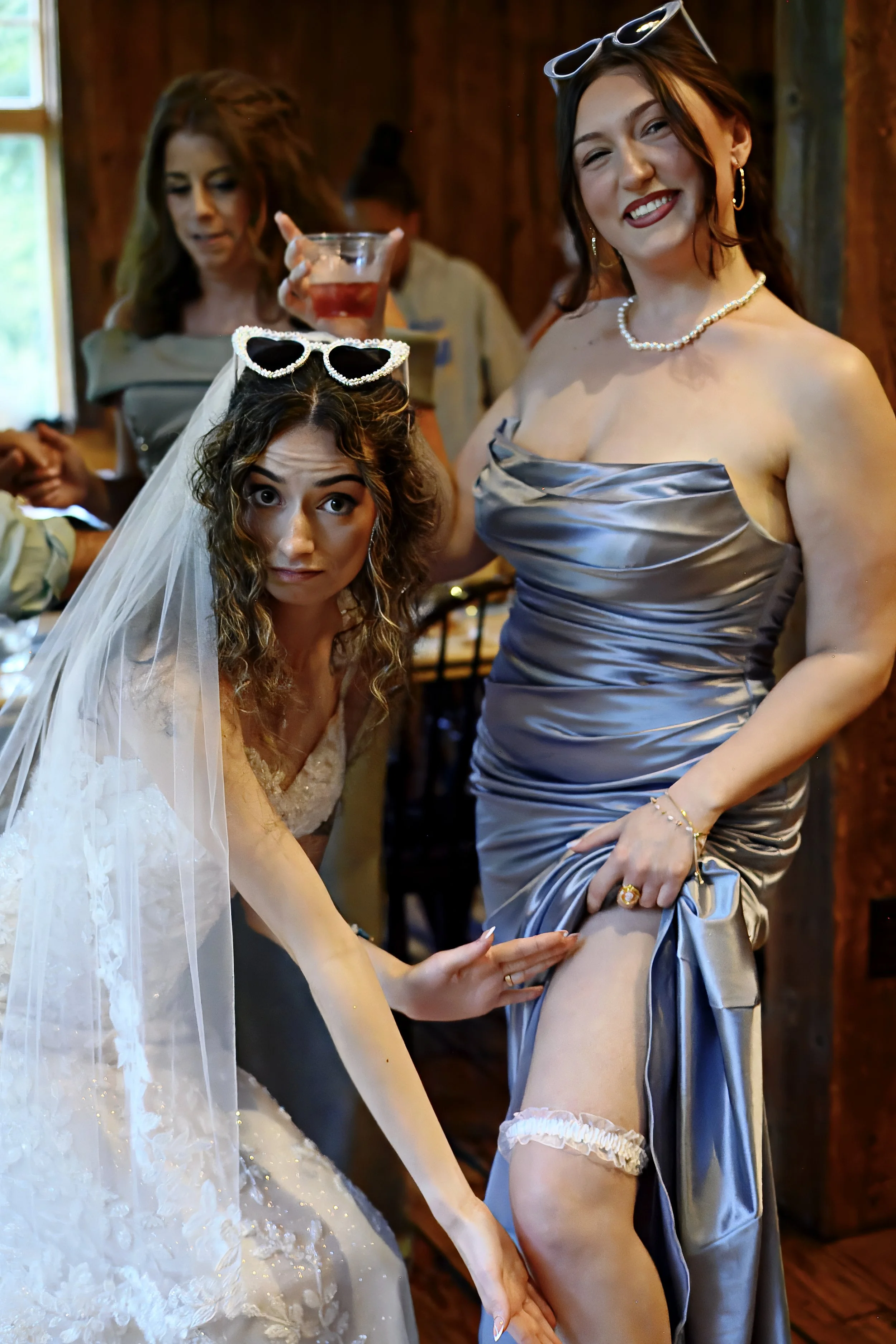 A woman in a wedding veil and dress kneels while another woman in a gray satin dress lifts her skirt, revealing a garter on her thigh. The woman in the gray dress is smiling and holding a glass of drink, with sunglasses on her head and jewelry. Other