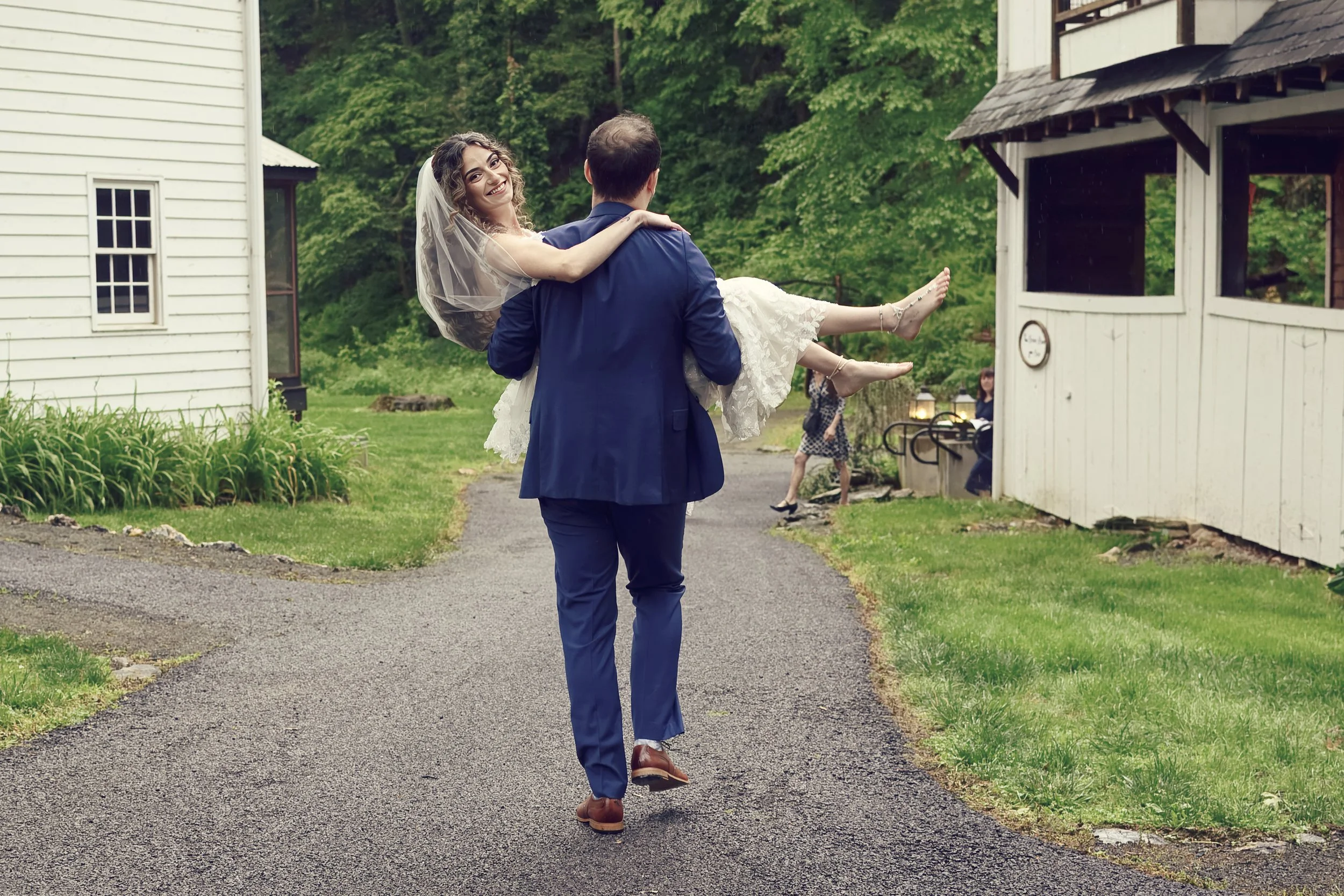 A man in a blue suit carrying a woman in a white wedding dress outdoors near houses, with trees and green foliage in the background.
