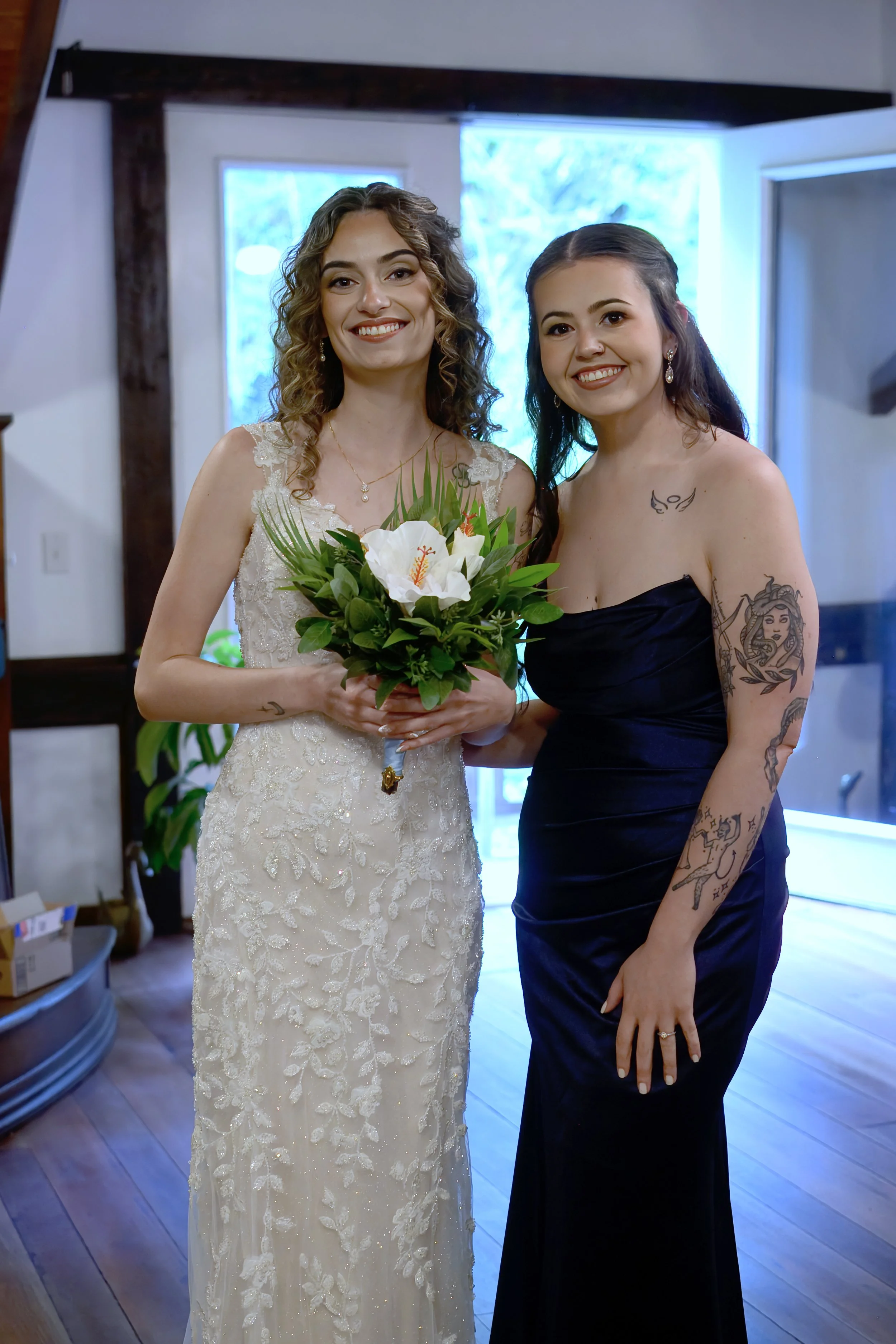 Two women smiling and posing for a photo indoors, one in a wedding dress holding a bouquet, and the other in a black strapless dress with tattoos on her arms and shoulder.
