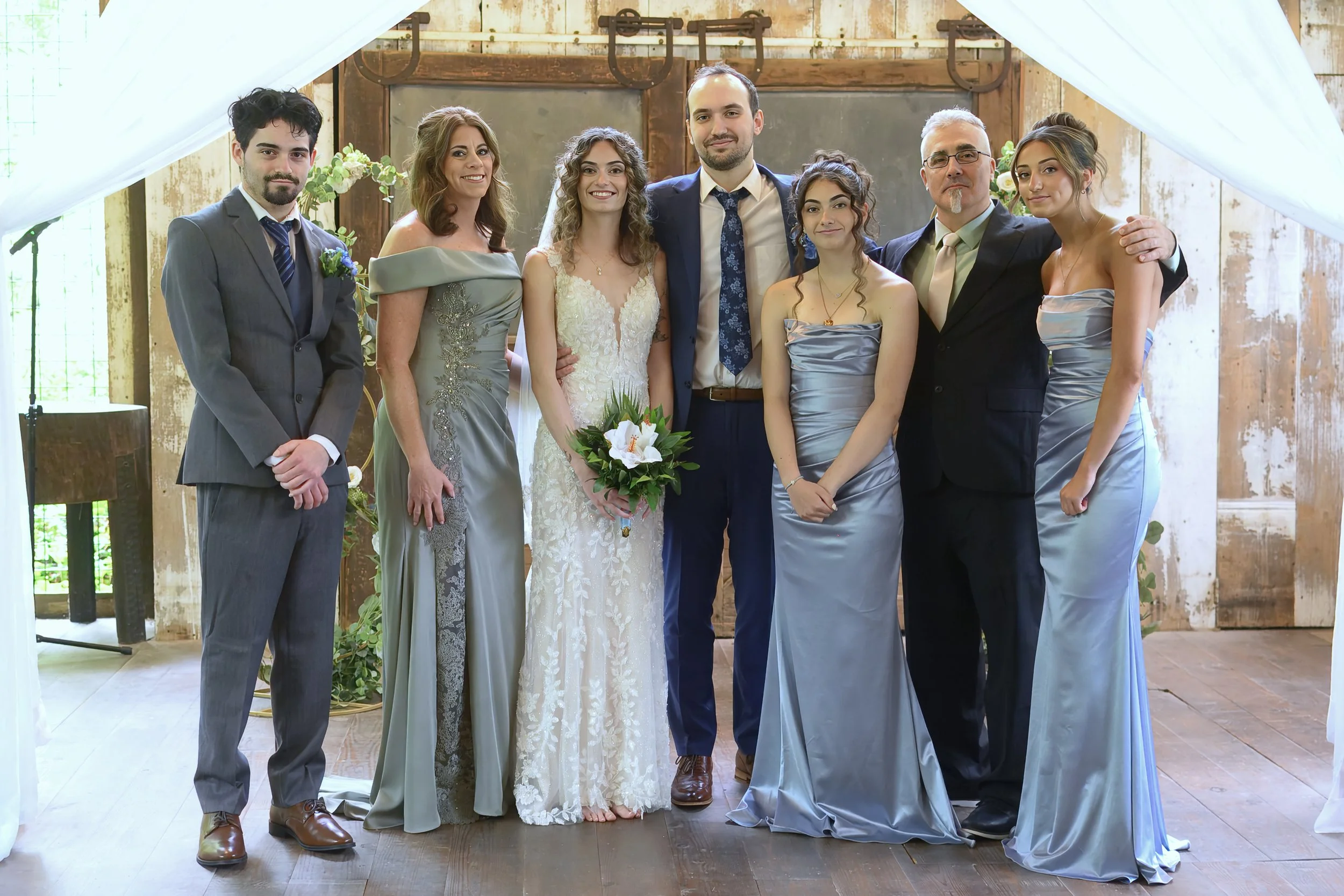A group of eight people, including a bride in a white wedding dress holding a bouquet, and others dressed in formal attire are gathered together and smiling at a wedding celebration inside a rustic venue with wooden walls and natural lighting.