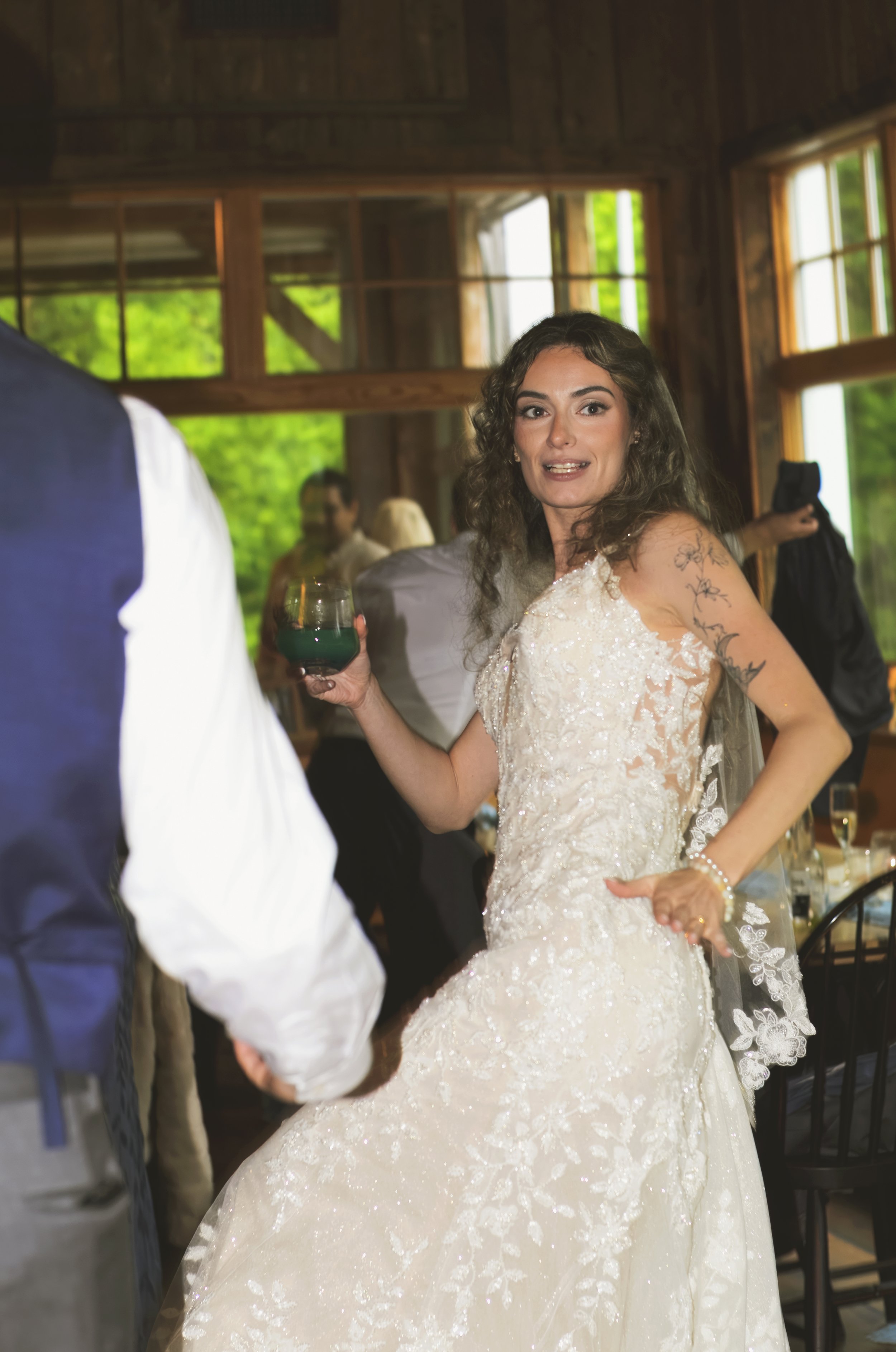Woman in a wedding dress holding a glass of blue drink, at a wedding reception with wooden interior and other guests in the background.