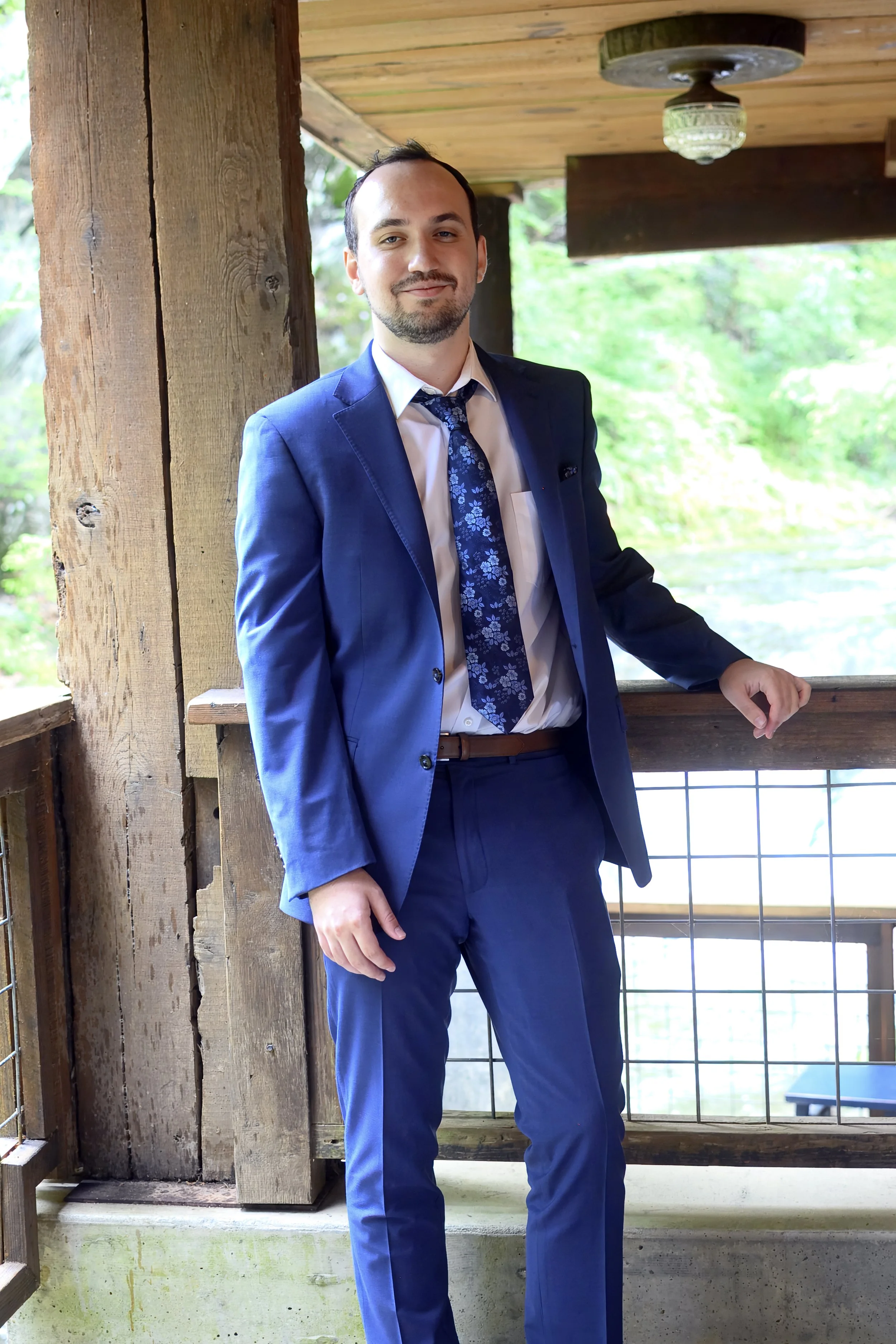 A man in a blue suit and dress shirt with a floral tie standing on a wooden balcony or porch with green trees in the background, smiling.