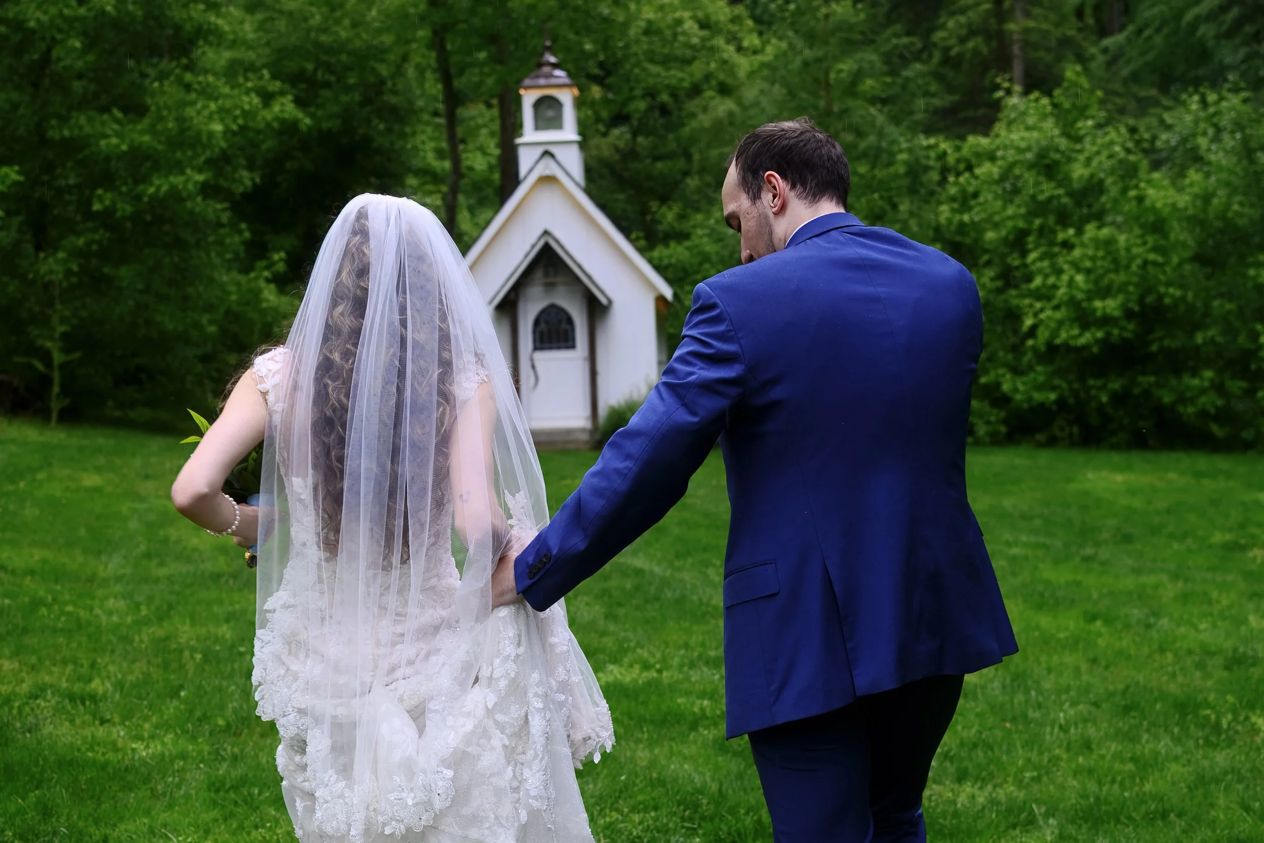 A bride in a white wedding dress with a veil and a groom in a blue suit holding hands outdoors near a small chapel surrounded by green trees.