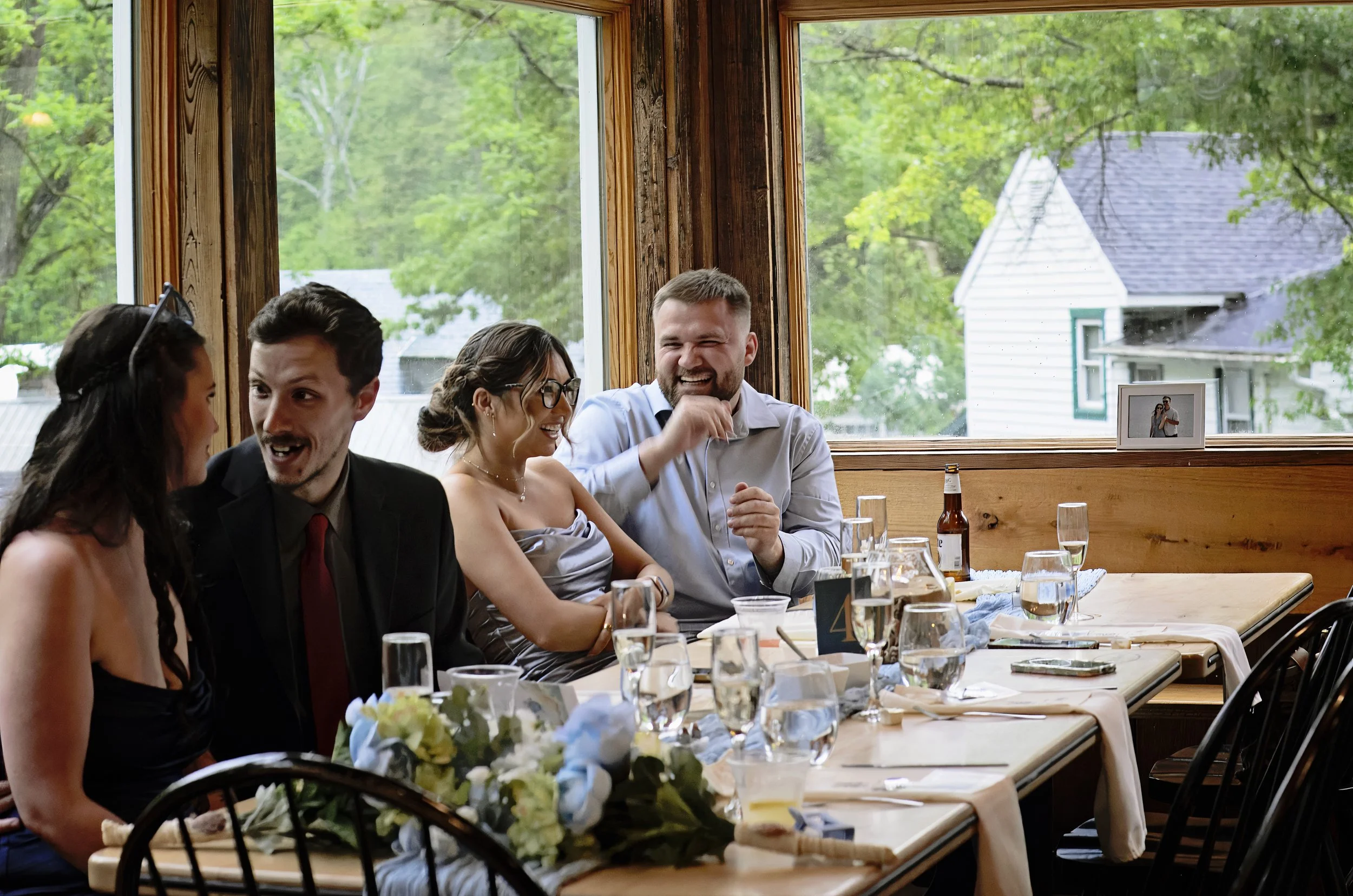 A group of people gathered around a table at a social event, smiling and interacting with each other inside a wooden-paneled room with large windows overlooking green trees and houses outside.