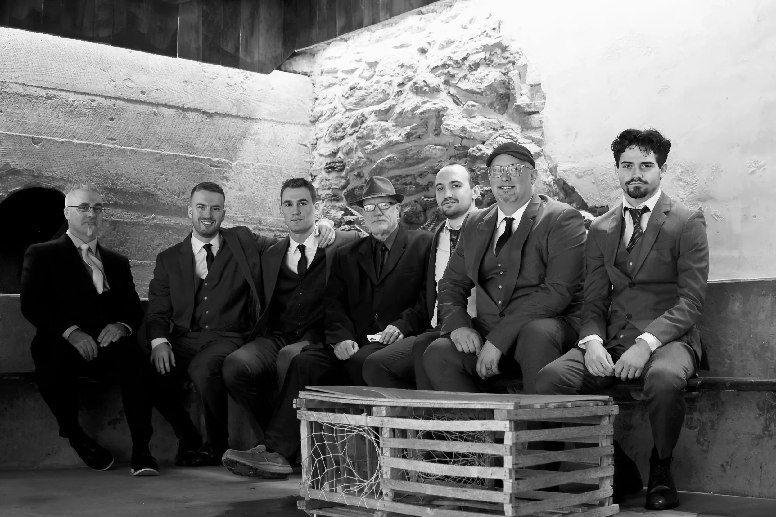 Black and white photo of seven men in suits sitting together in front of a stone and white wall, with a wooden crate with a fishing net underneath.