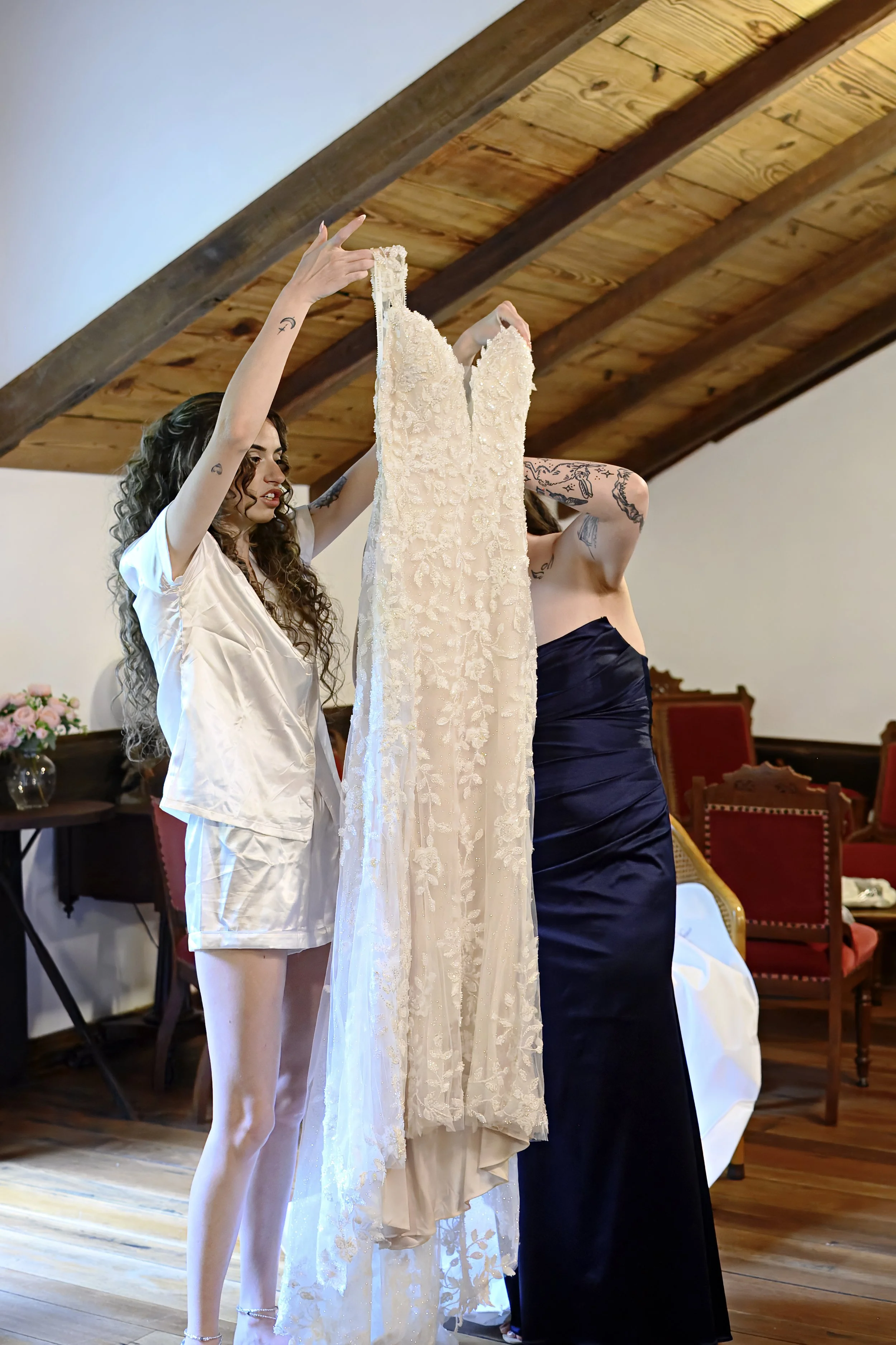 Two women in elegant dresses are holding a cream-colored, embroidered wedding dress, preparing for a wedding ceremony in a room with wooden ceiling beams and chairs.