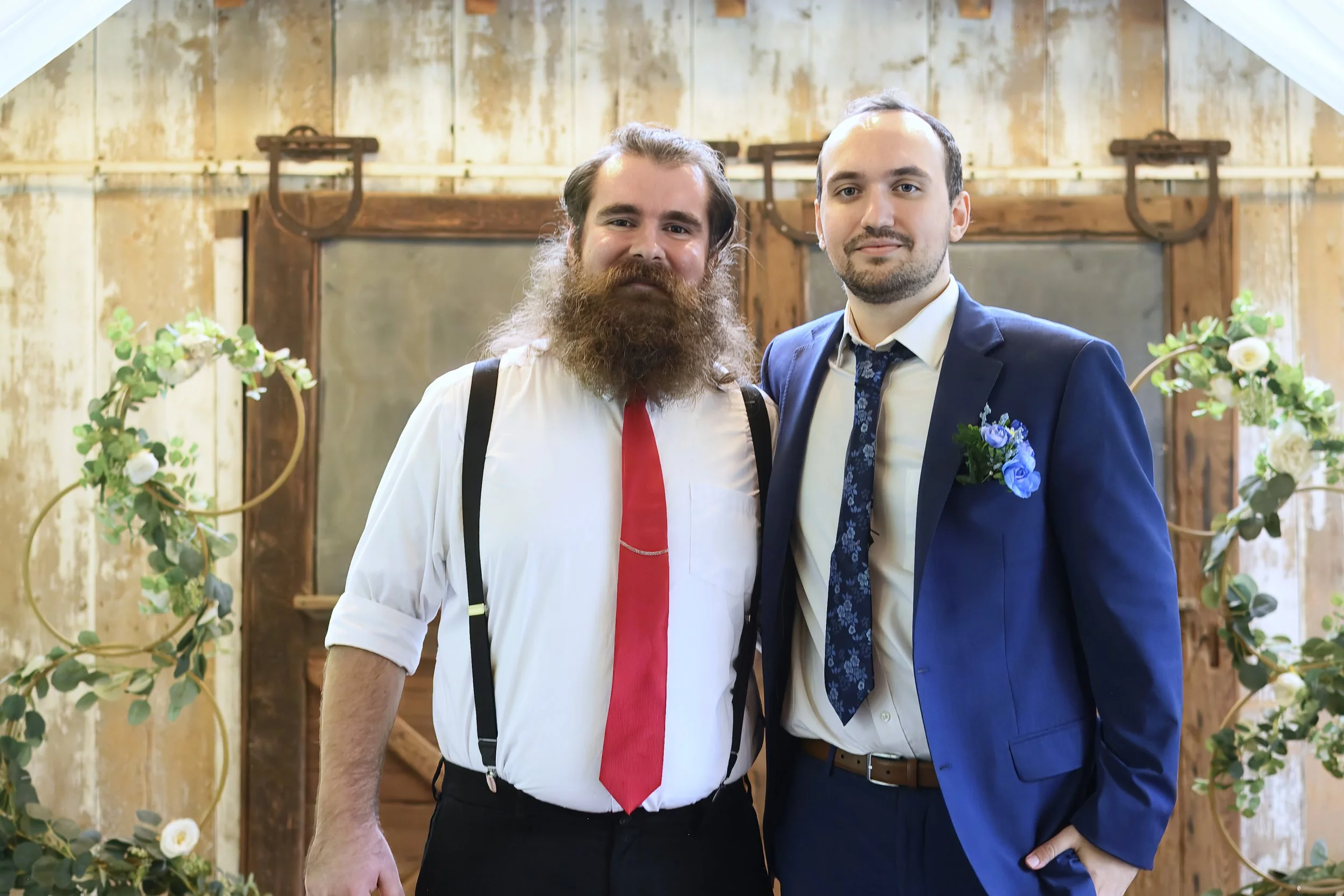 Two men posing at a rustic wedding or event venue, one with a beard and suspenders, the other in a suit with boutonniere, standing close together with floral decorations around them.