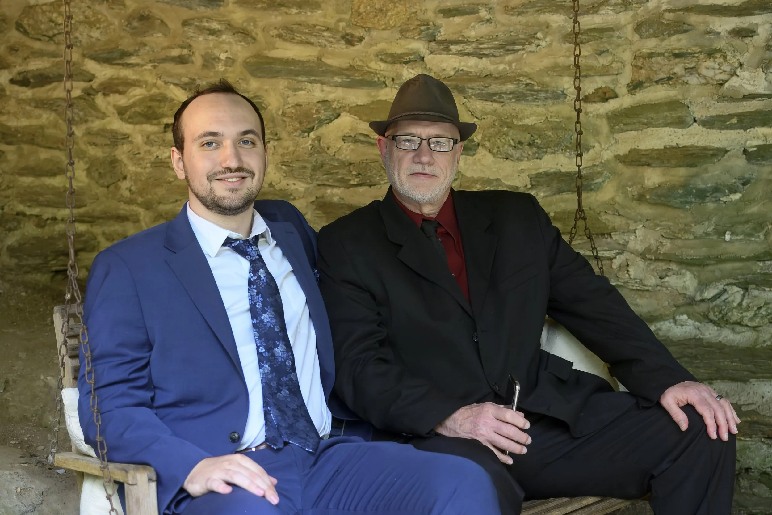 Two men sit on a wooden swing with metal chains against a stone wall background. One man is young, wearing a blue suit and patterned tie, and is smiling; the other is older, wearing glasses, a dark suit, a red shirt, and a hat, with a serious express