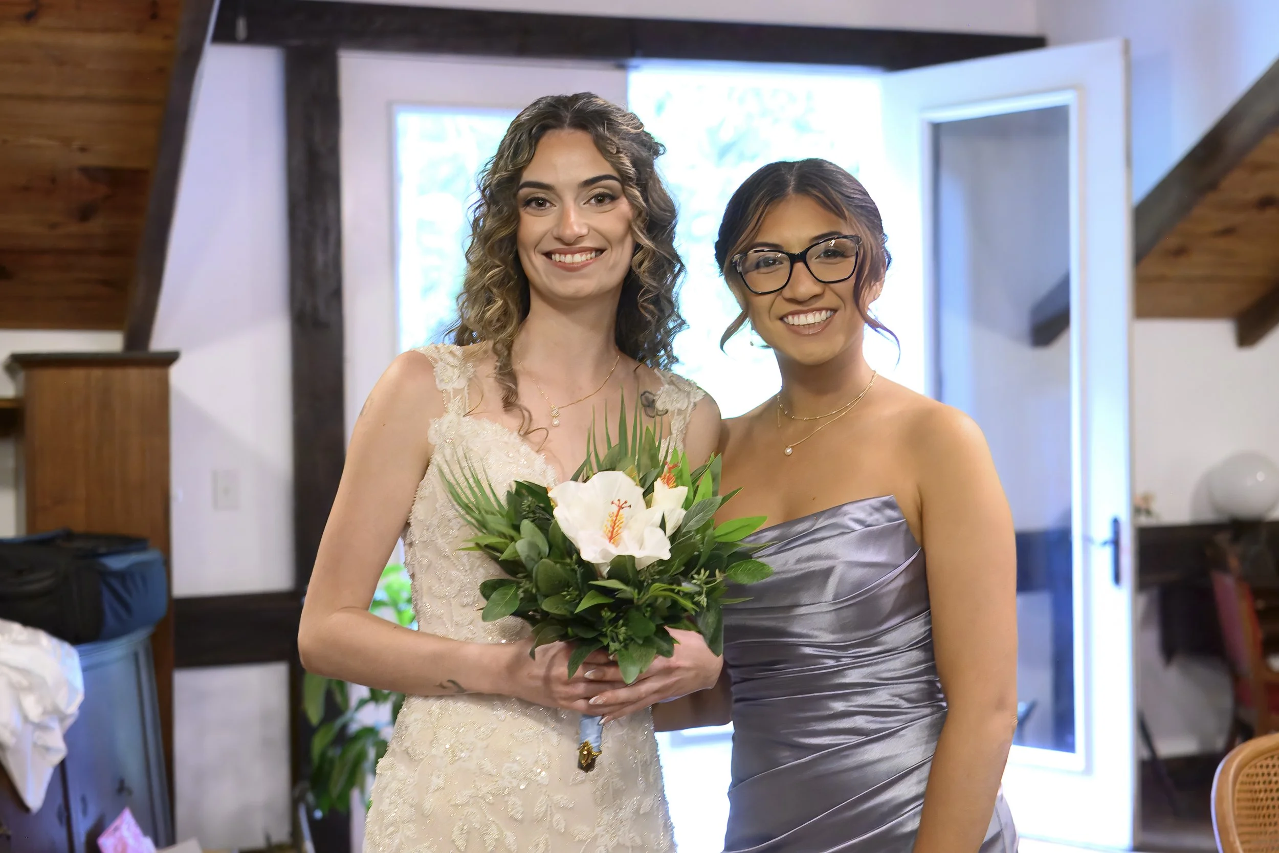 Two women at a wedding, one in a wedding dress holding a bouquet, and the other in a silver gown. They are smiling inside a room with wooden beams and large windows.