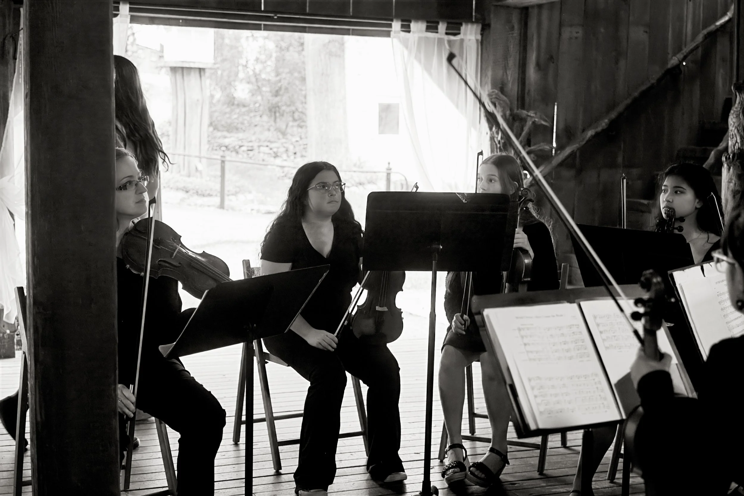 Group of women musicians sitting with violins and sheet music in a rustic wooden room, possibly rehearsing or performing.
