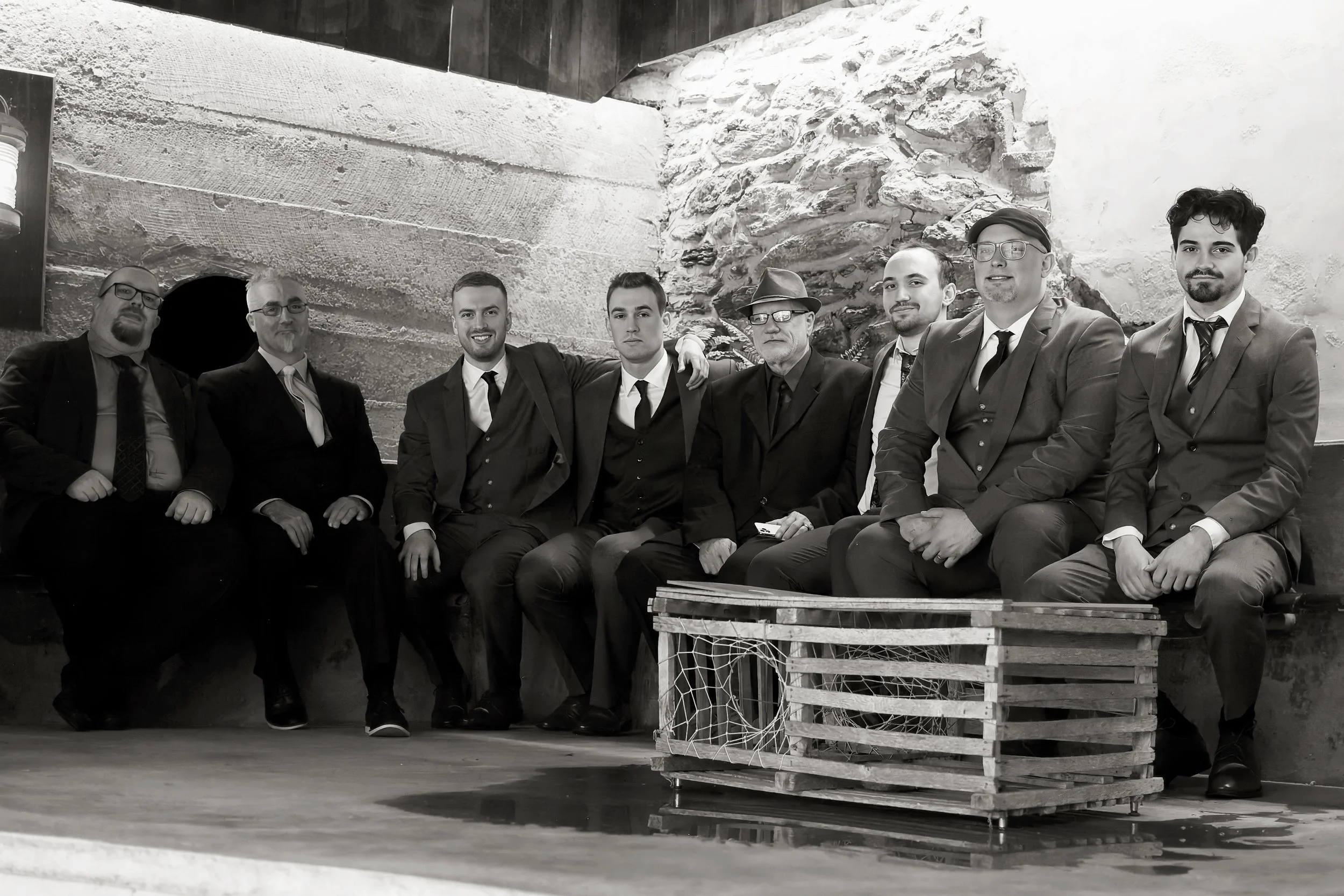 A black and white photo of a group of nine men dressed in suits, sitting and standing in a rustic room with wooden and stone walls. They are posing for a picture, some with arms around each other. There is a wooden pallet and some fishing nets in the